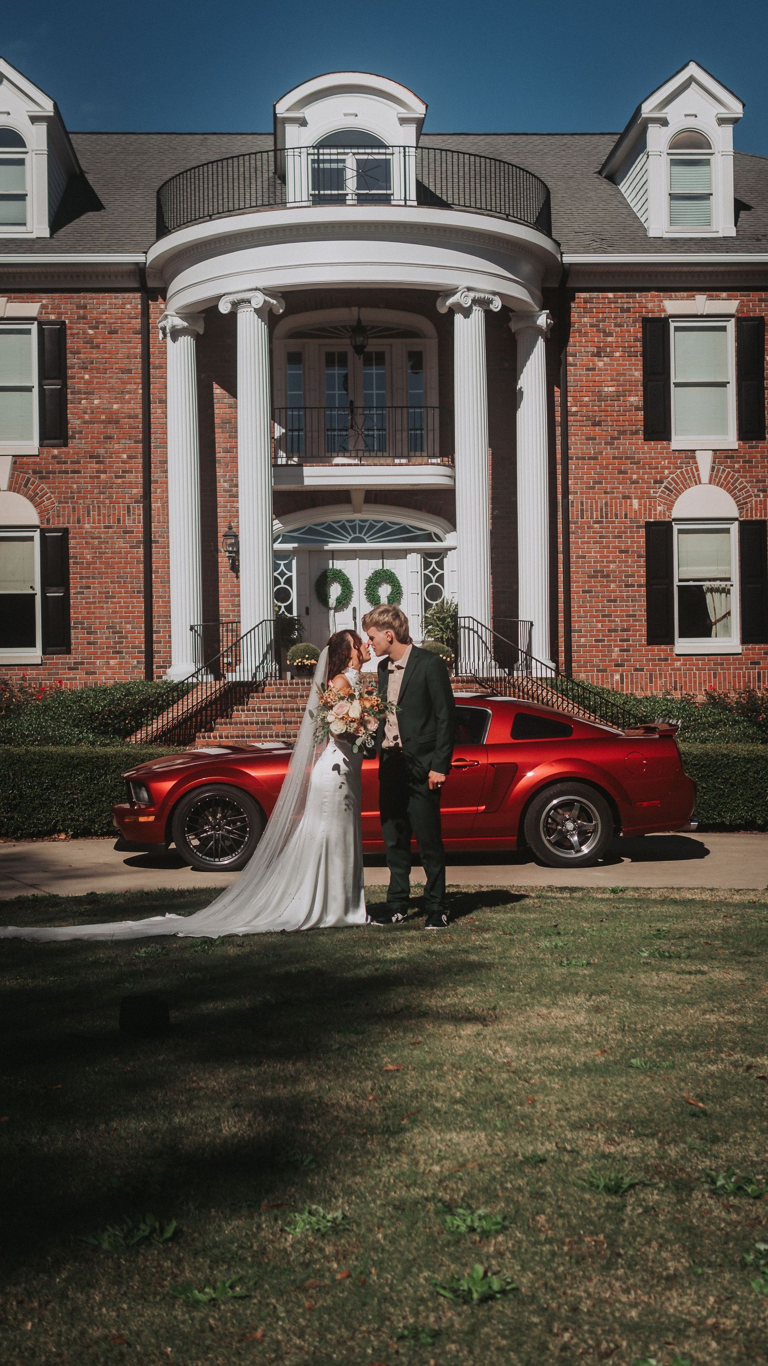 Bride and groom kissing in front of a red Mustang and elegant brick mansion with white columns on their wedding day.
