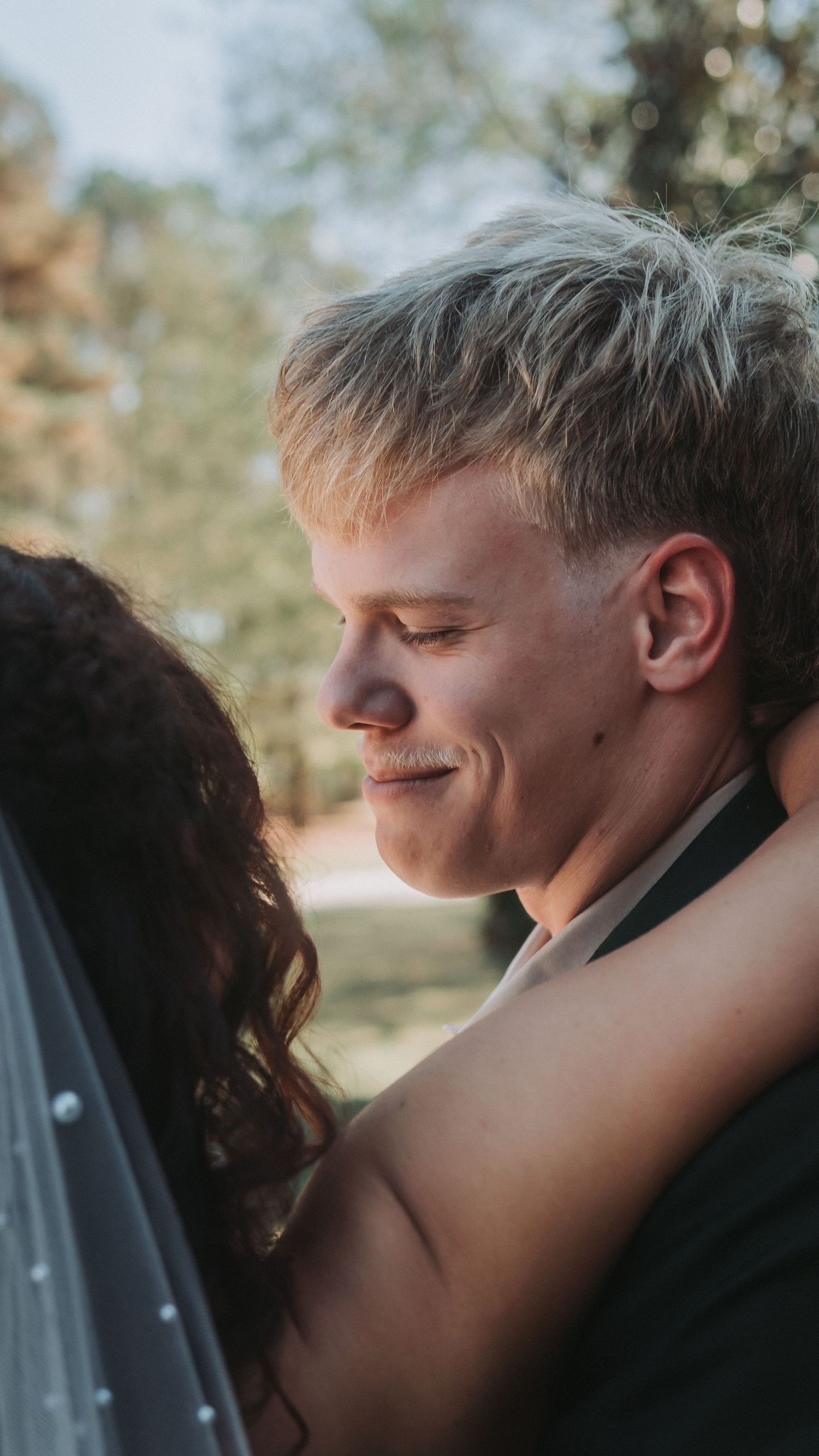 Smiling groom with blonde hair embraces bride in an intimate outdoor wedding moment.