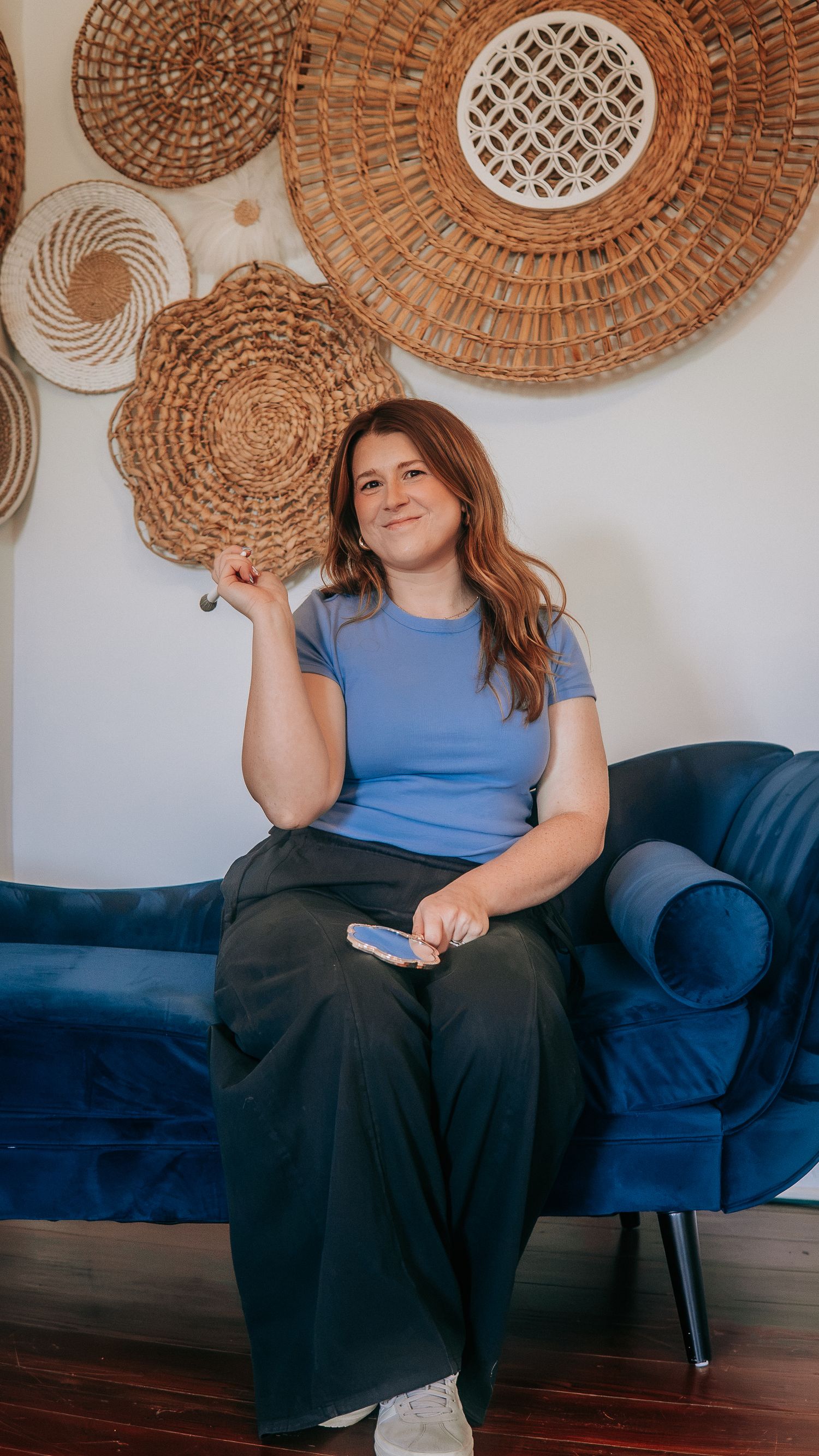 Woman in blue t-shirt and black wide-leg pants sitting on navy velvet sofa with woven basket wall decor behind her.