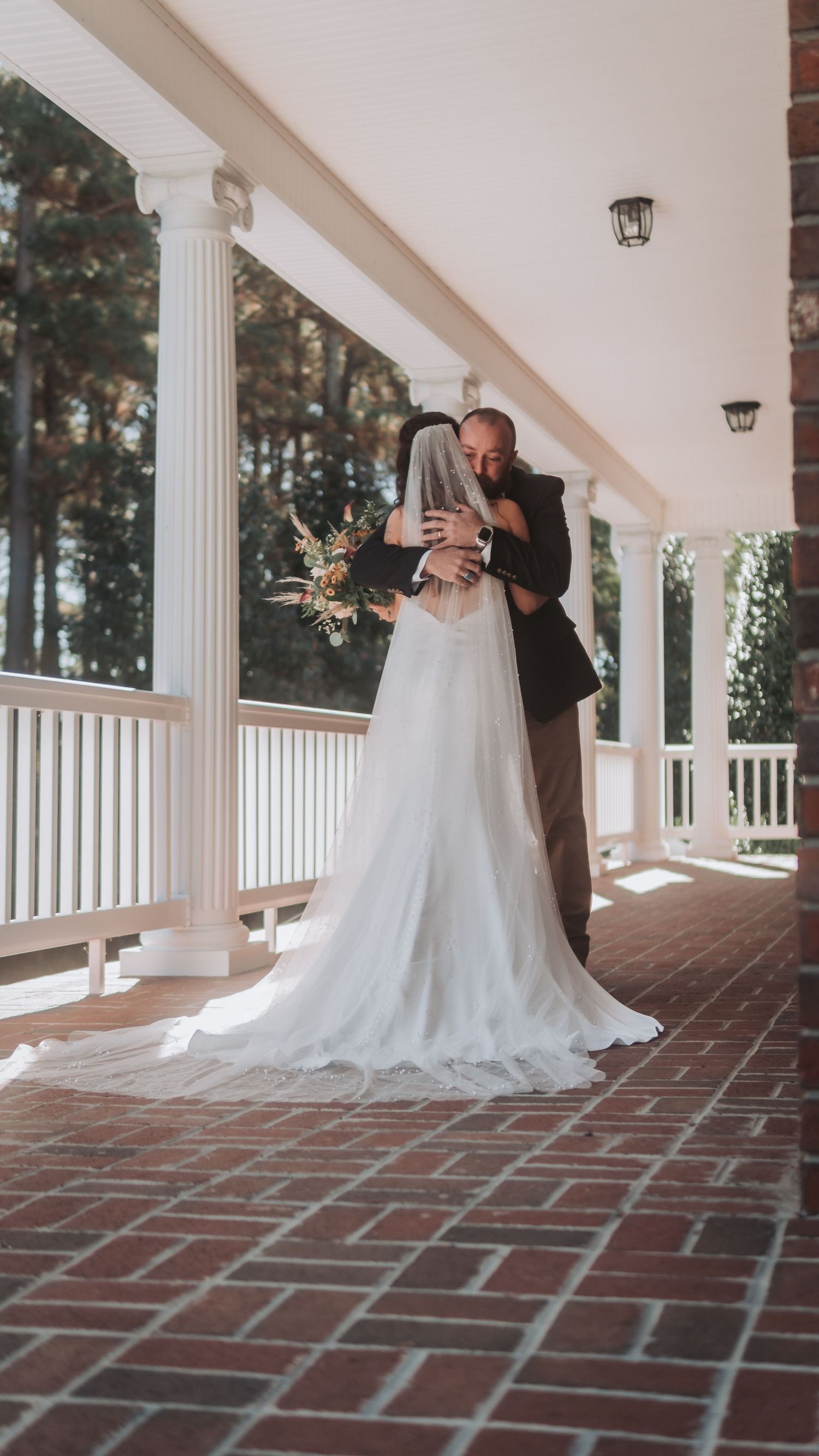 Bride and groom share a romantic embrace on a classic white columned porch, bride wearing a flowing white gown with veil and bouquet.