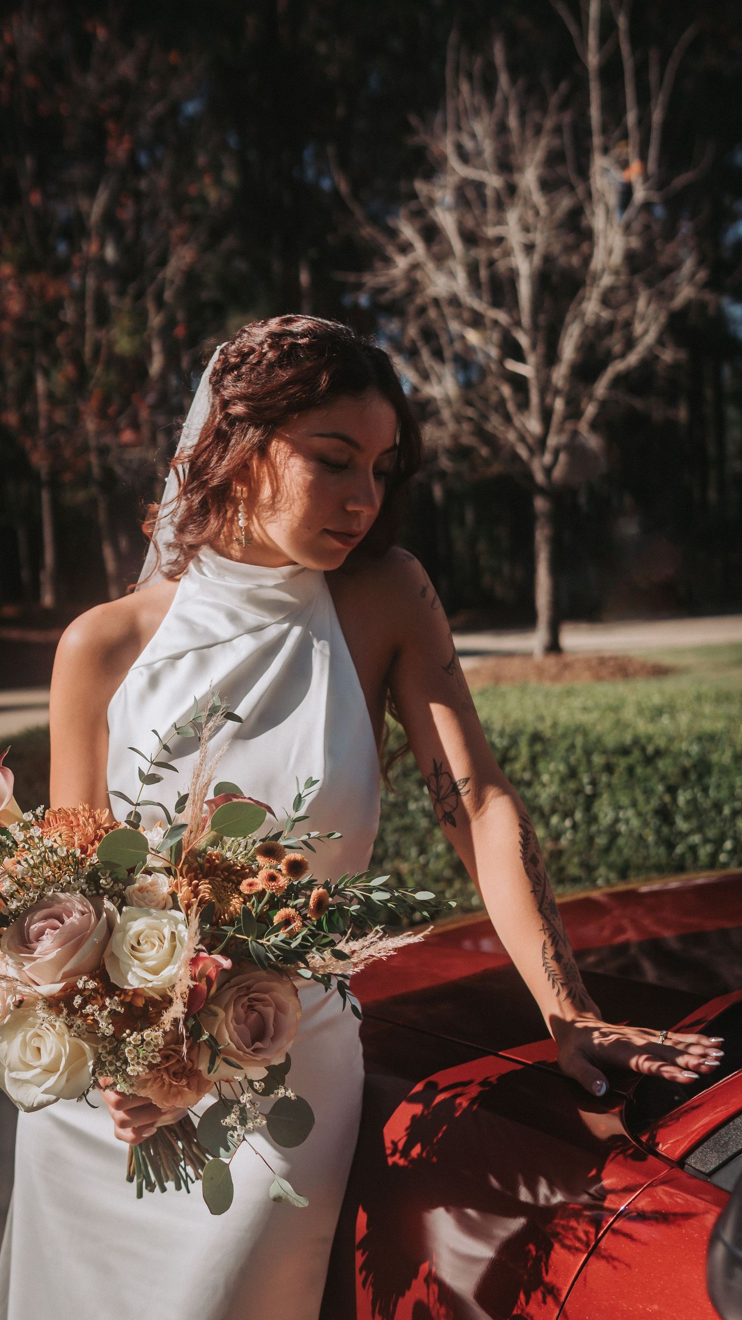 Bride in white halter gown holding autumn bouquet of roses beside a red car outdoors.