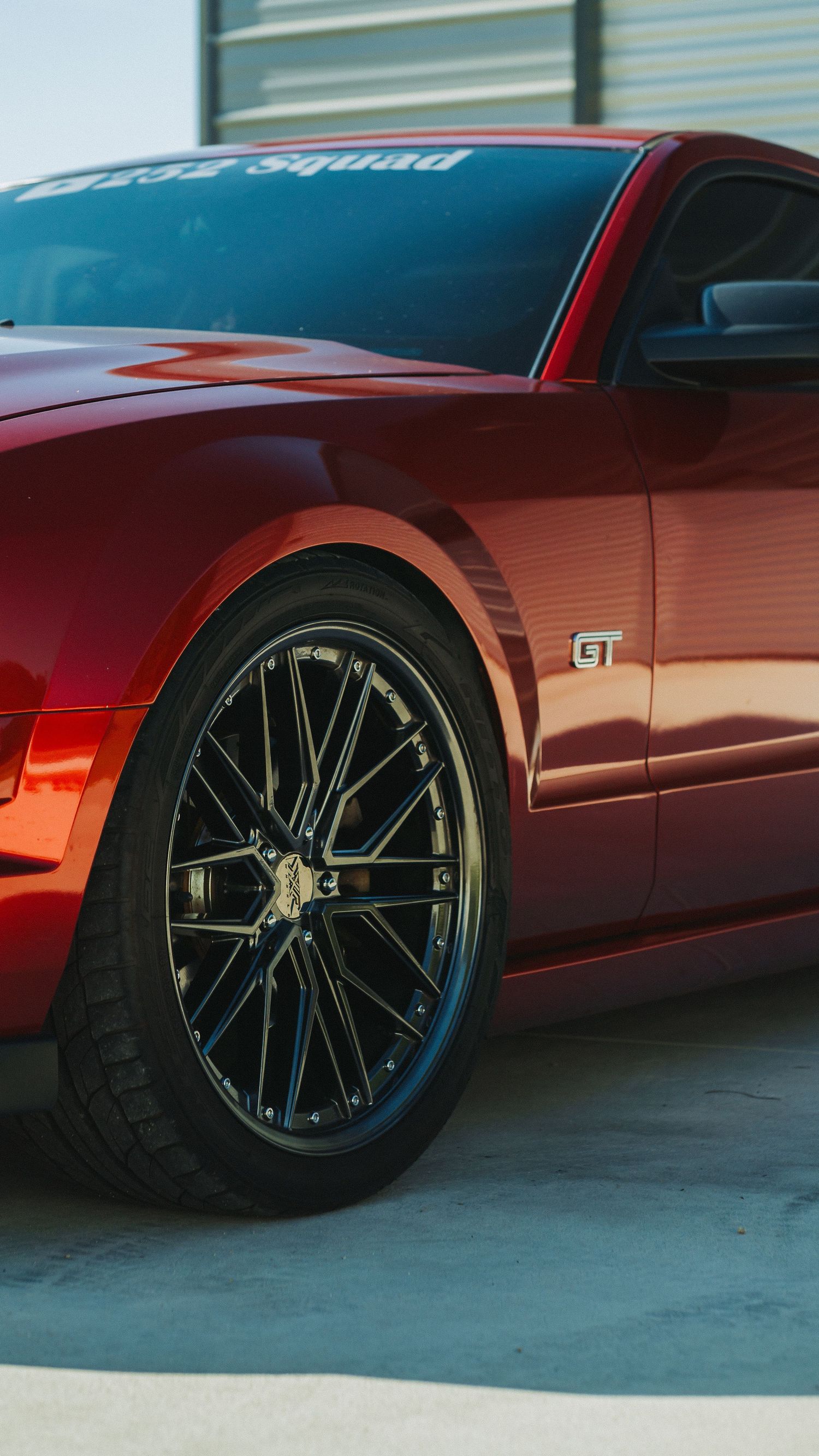 Red Ford Mustang GT with black multi-spoke wheels parked outdoors, featuring a GT badge and custom windshield banner.