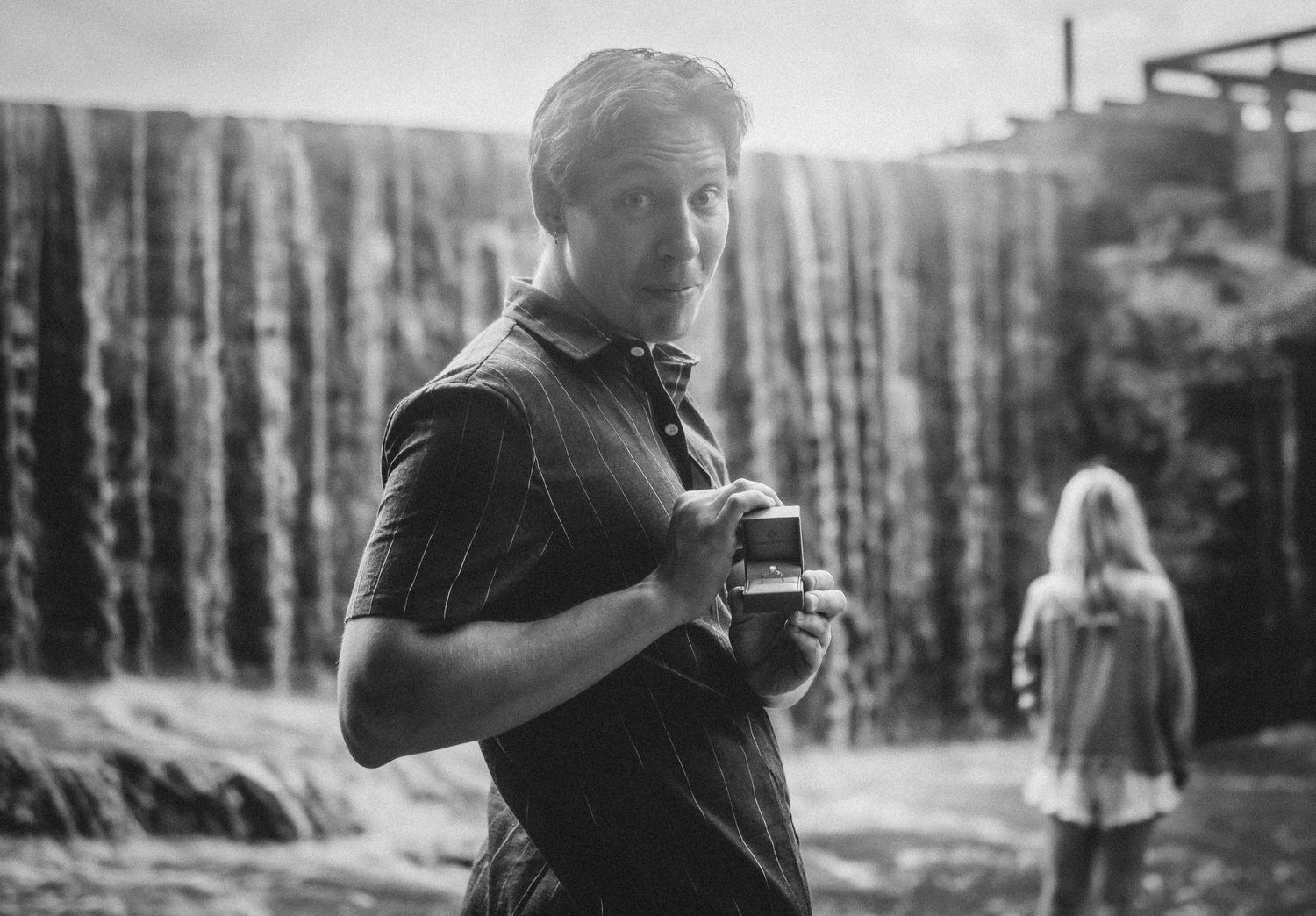 Black and white photo of a man holding a drink near a waterfall with a woman standing in the background.