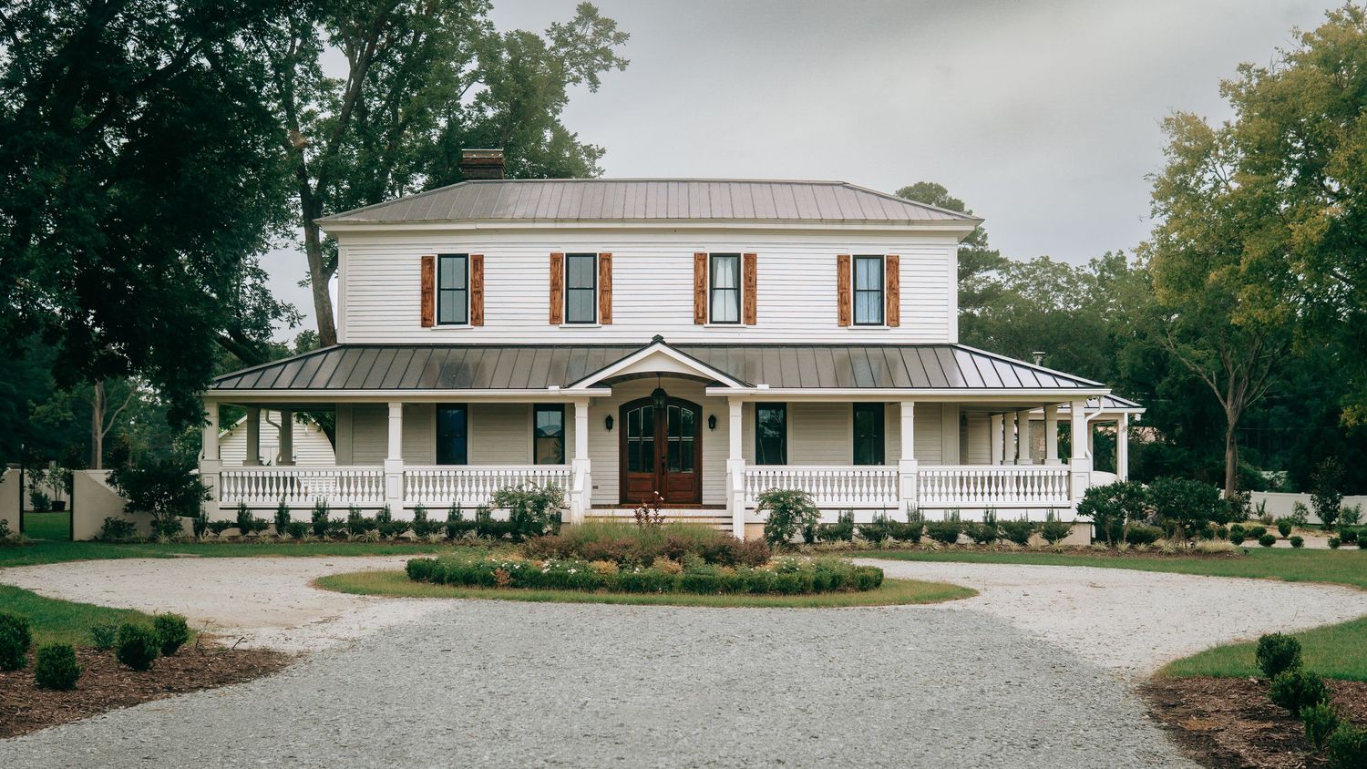 White farmhouse with metal roof, wrap-around porch, red shutters, and circular gravel driveway surrounded by lush trees.