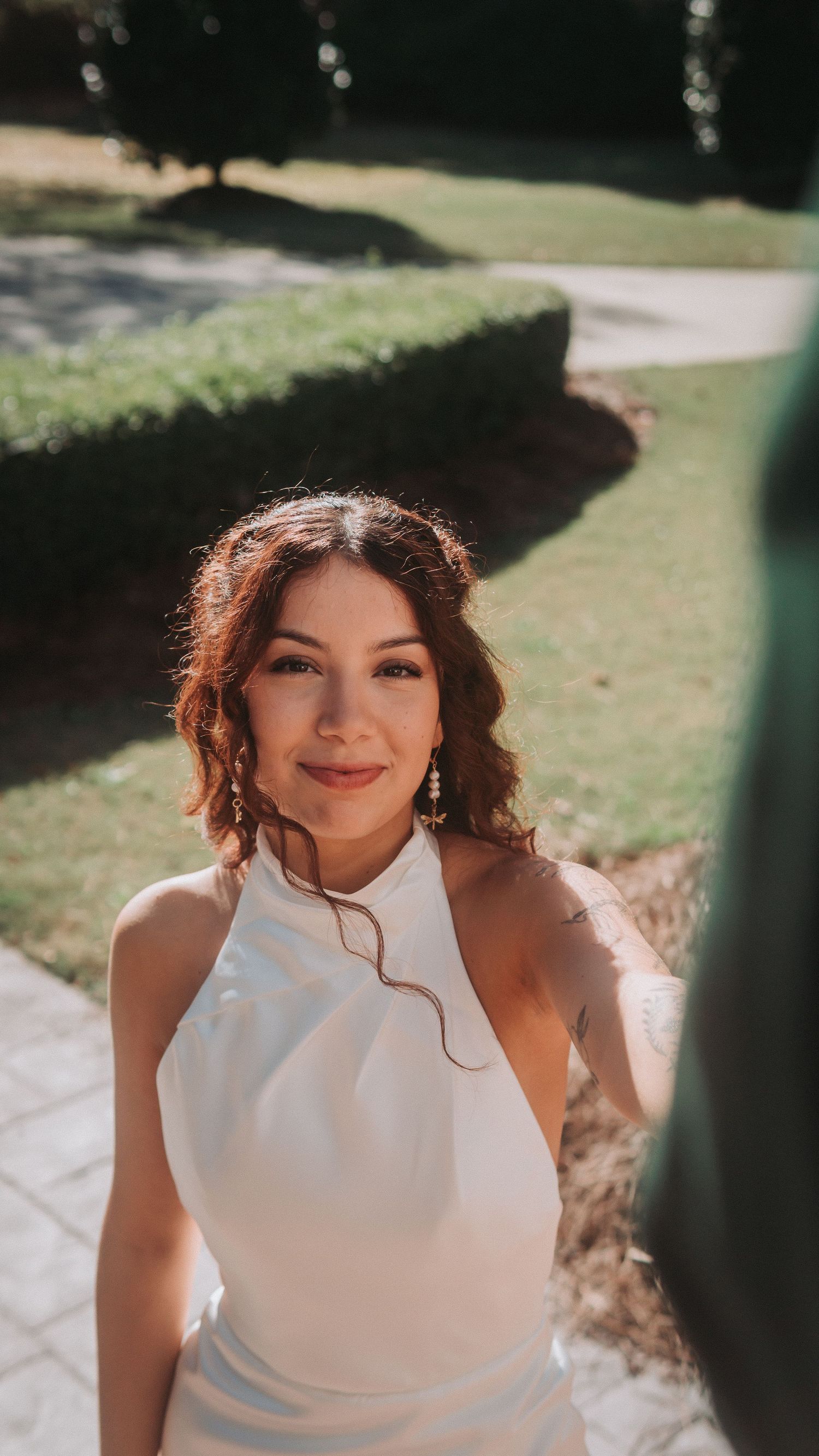 Woman in white halter dress smiling outdoors in sunlit garden setting with green hedges.