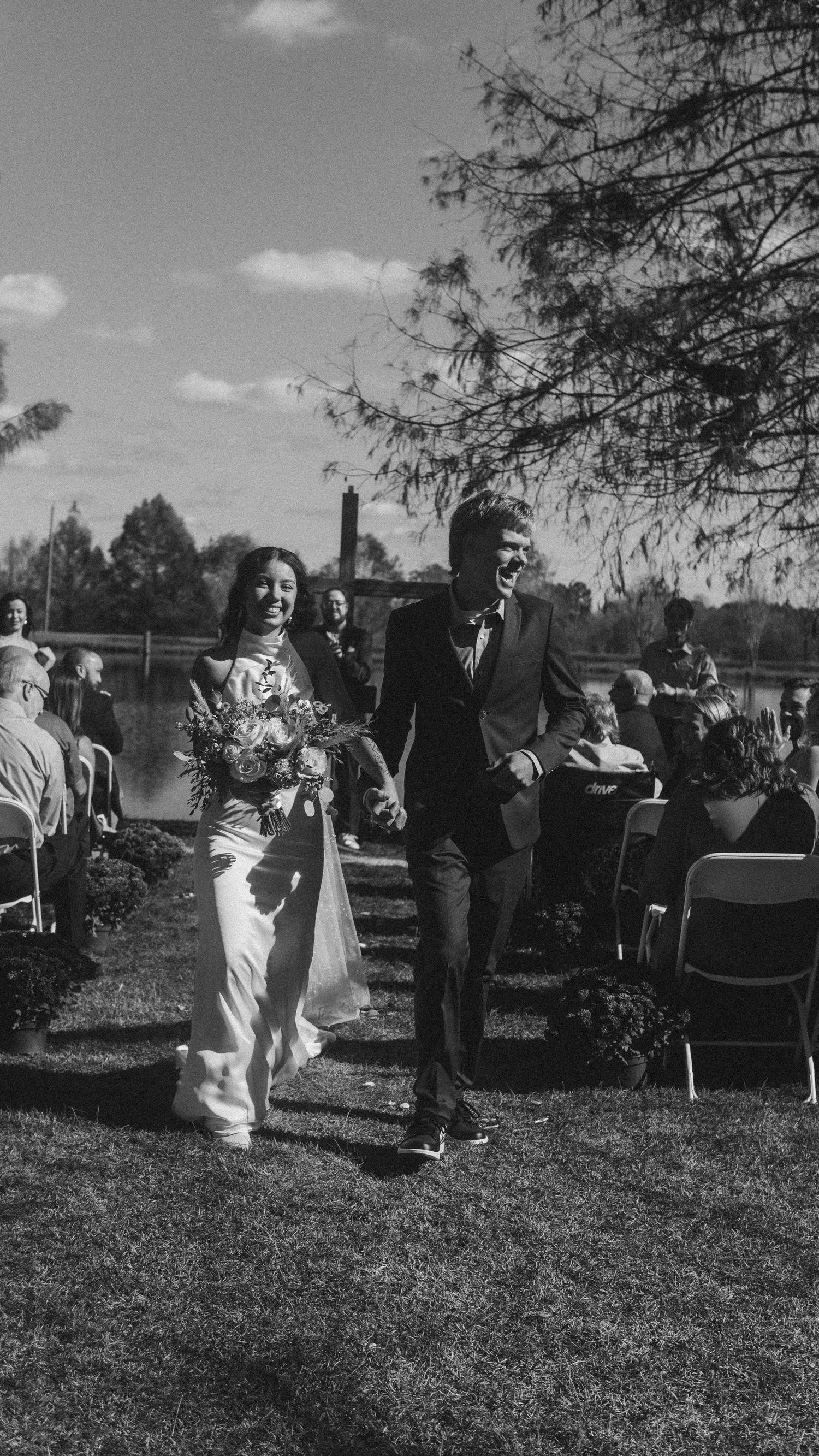 Black and white photo of a joyful bride and groom walking down the aisle at an outdoor wedding ceremony.