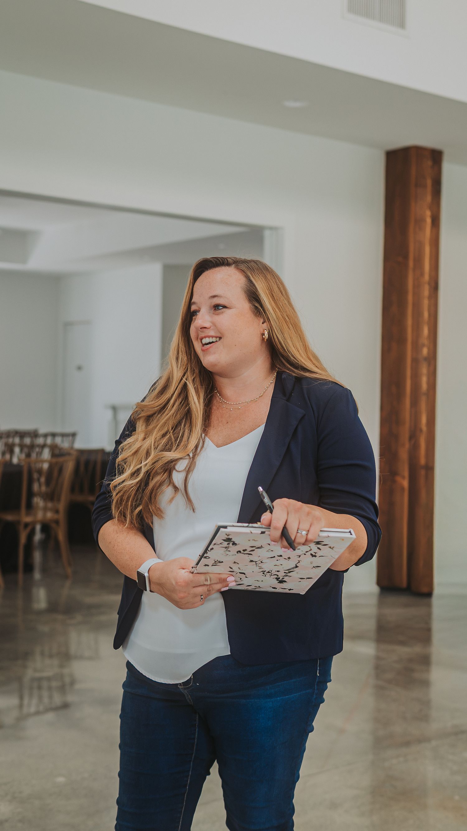Smiling woman in navy blazer holds clipboard and pen while standing in bright modern event venue space.