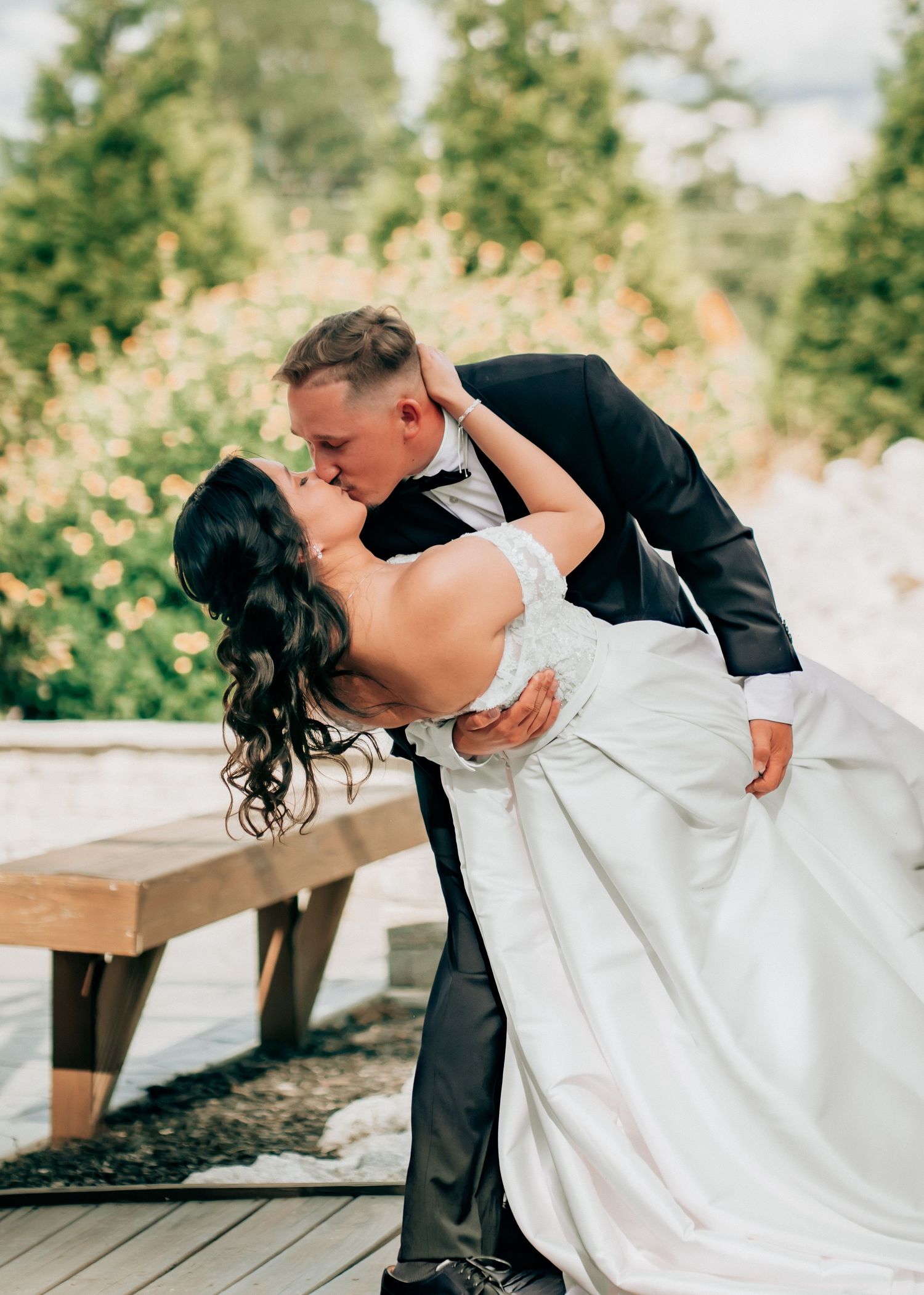 A groom dips his bride for a romantic kiss outdoors, she wears a white gown as he holds her in a dark suit.