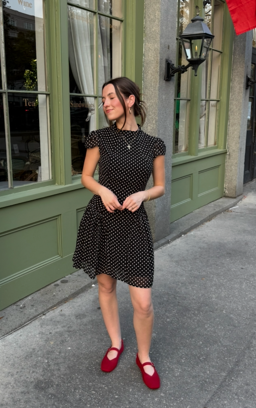 Woman in black polka dot mini dress and red Mary Jane flats standing on sidewalk outside green storefront.