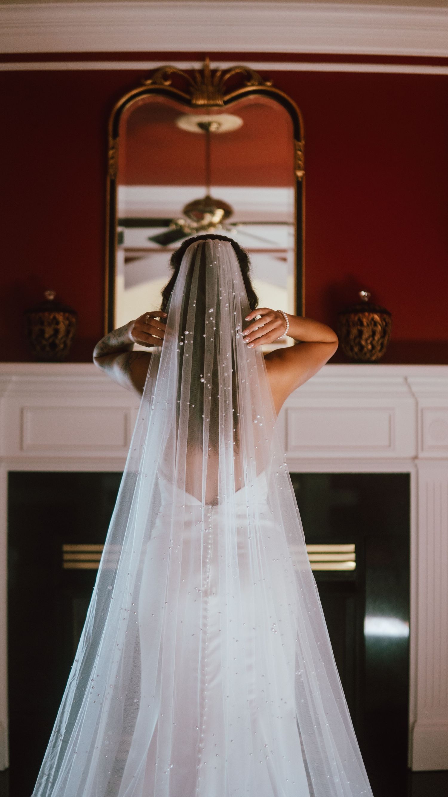 Bride in white fitted gown adjusts her long pearl veil while standing before an ornate gold mirror.
