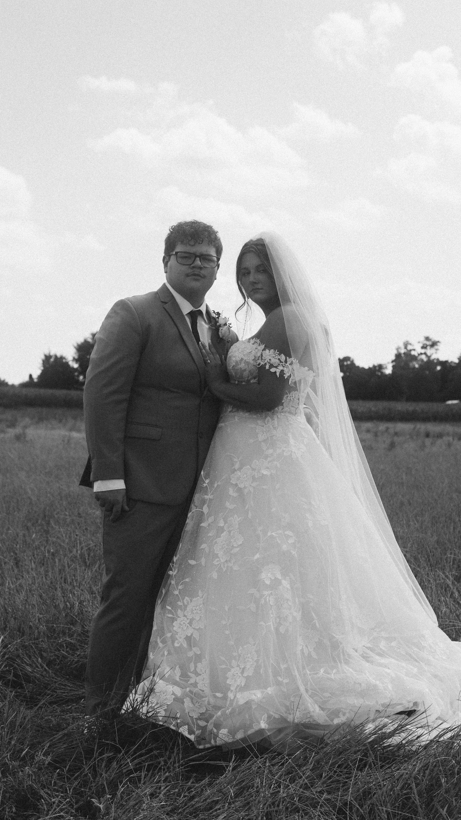 Black and white photo of a bride and groom posing in a field, bride in lace gown with veil, groom in suit.