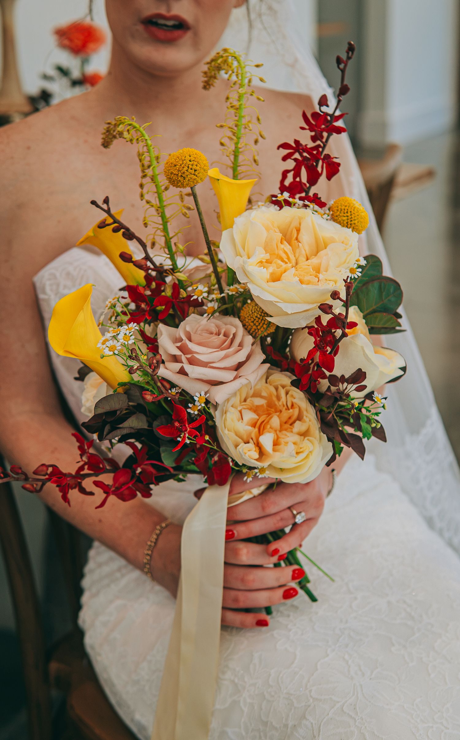 Bride in white lace gown holding a vibrant bouquet with yellow calla lilies, peach roses, red orchids, and golden accents.