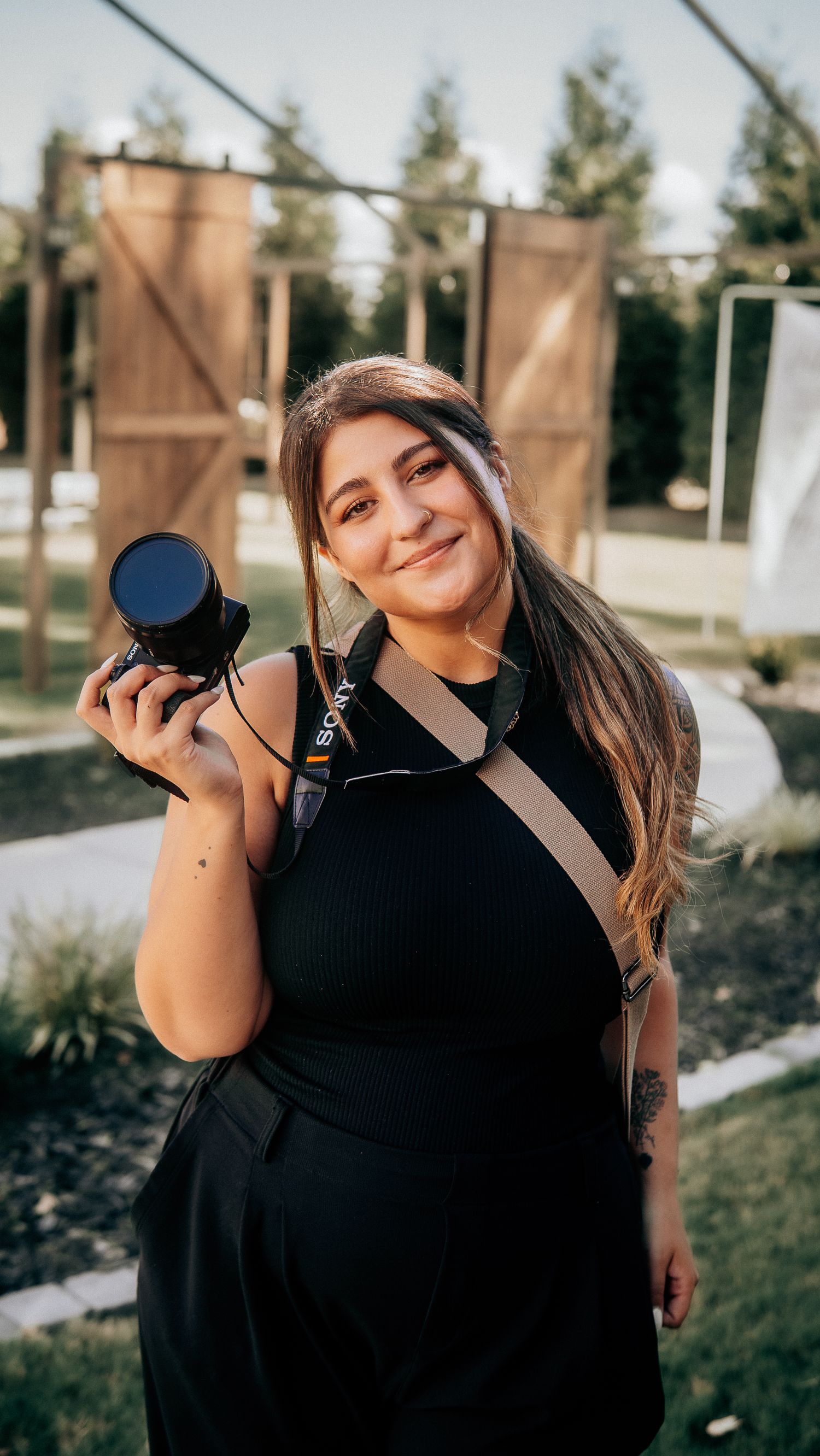 Smiling woman in black dress holding a camera with a Sony strap outdoors near wooden barn doors.