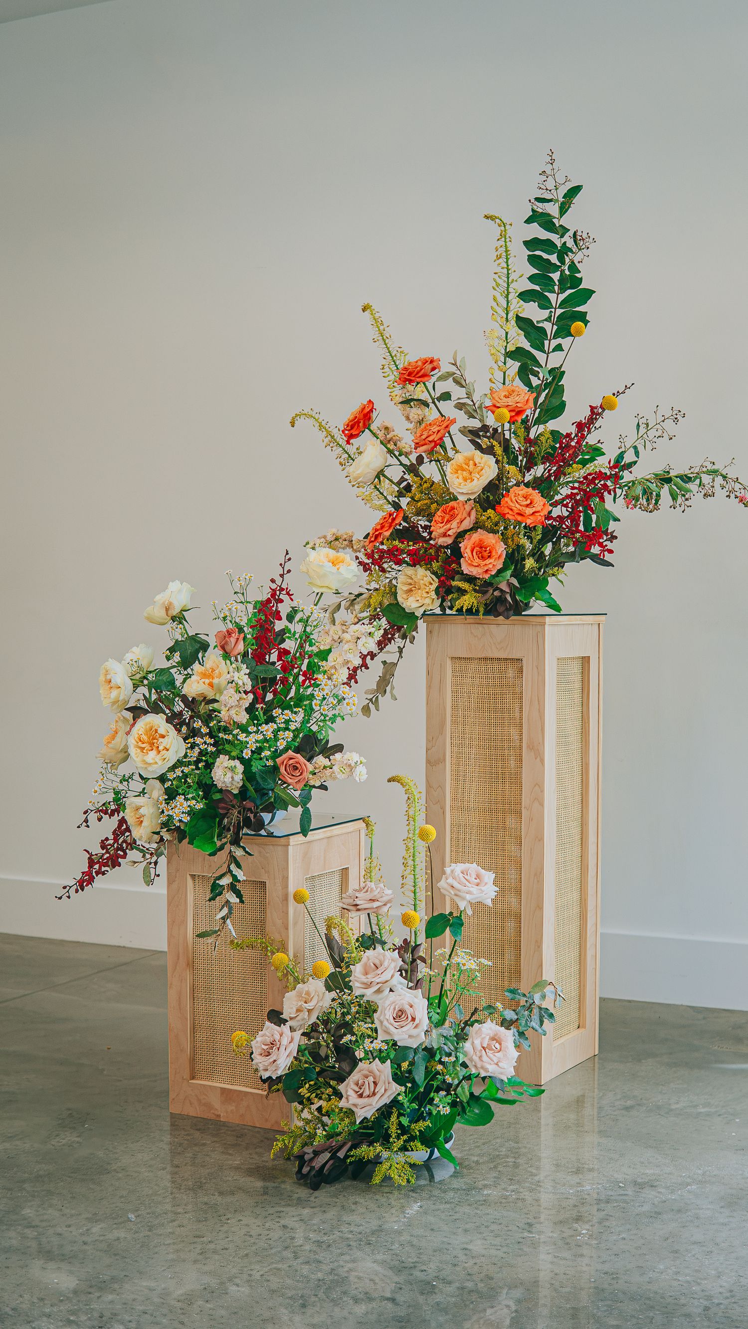 Three floral arrangements with roses and greenery displayed on wooden pedestals in a bright gallery space.