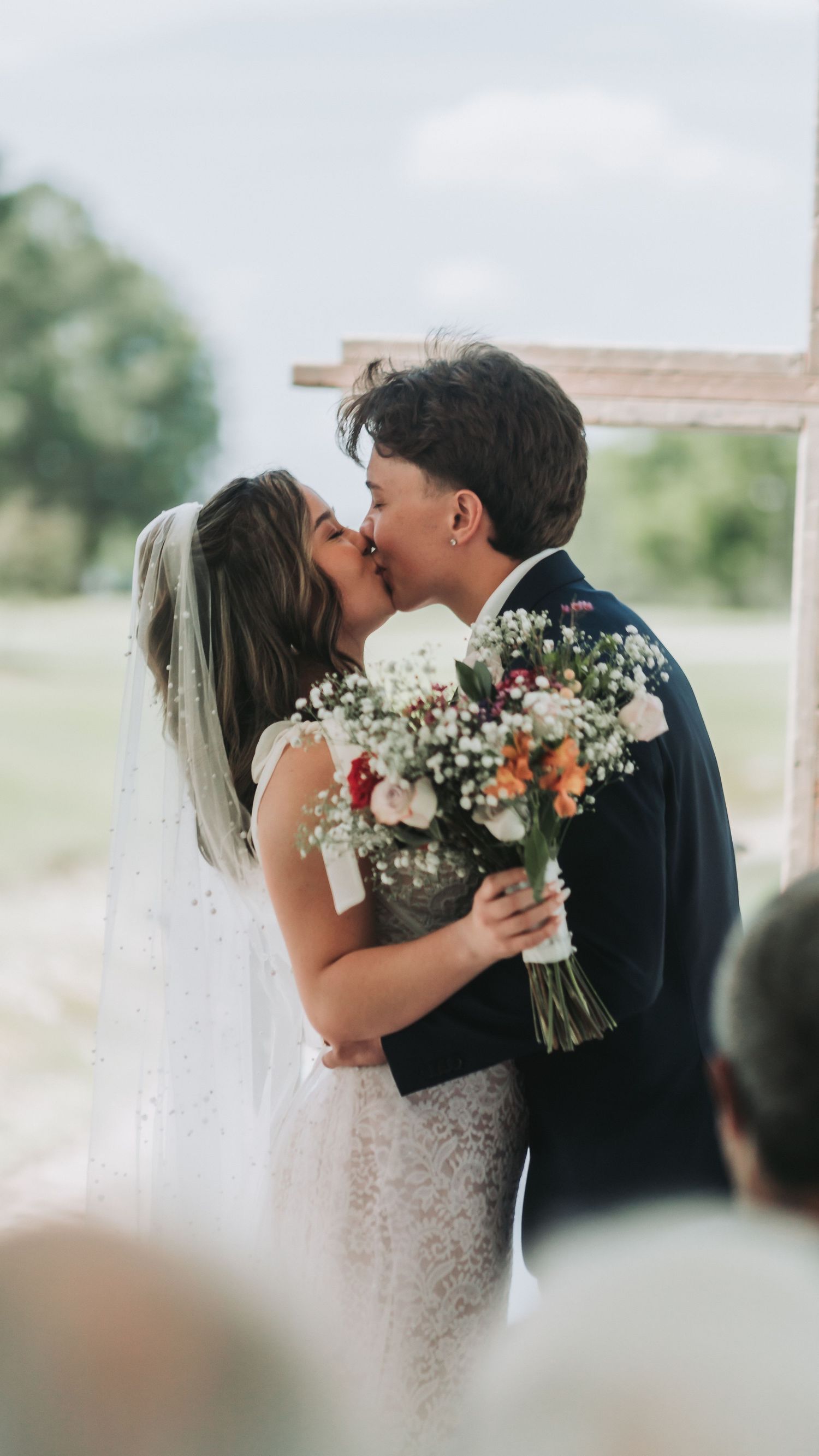 Bride and groom share first kiss at outdoor wedding ceremony, bride holding colorful bouquet with roses and baby's breath.