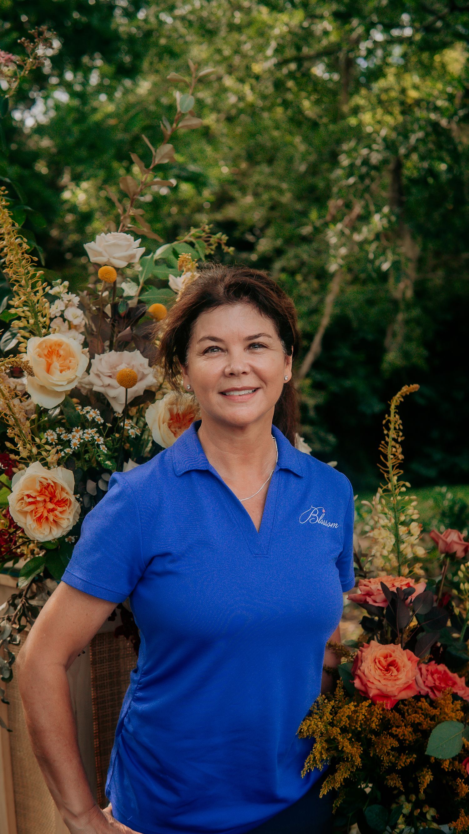 Woman in blue polo shirt smiling outdoors surrounded by colorful floral arrangements with lush green trees in background.