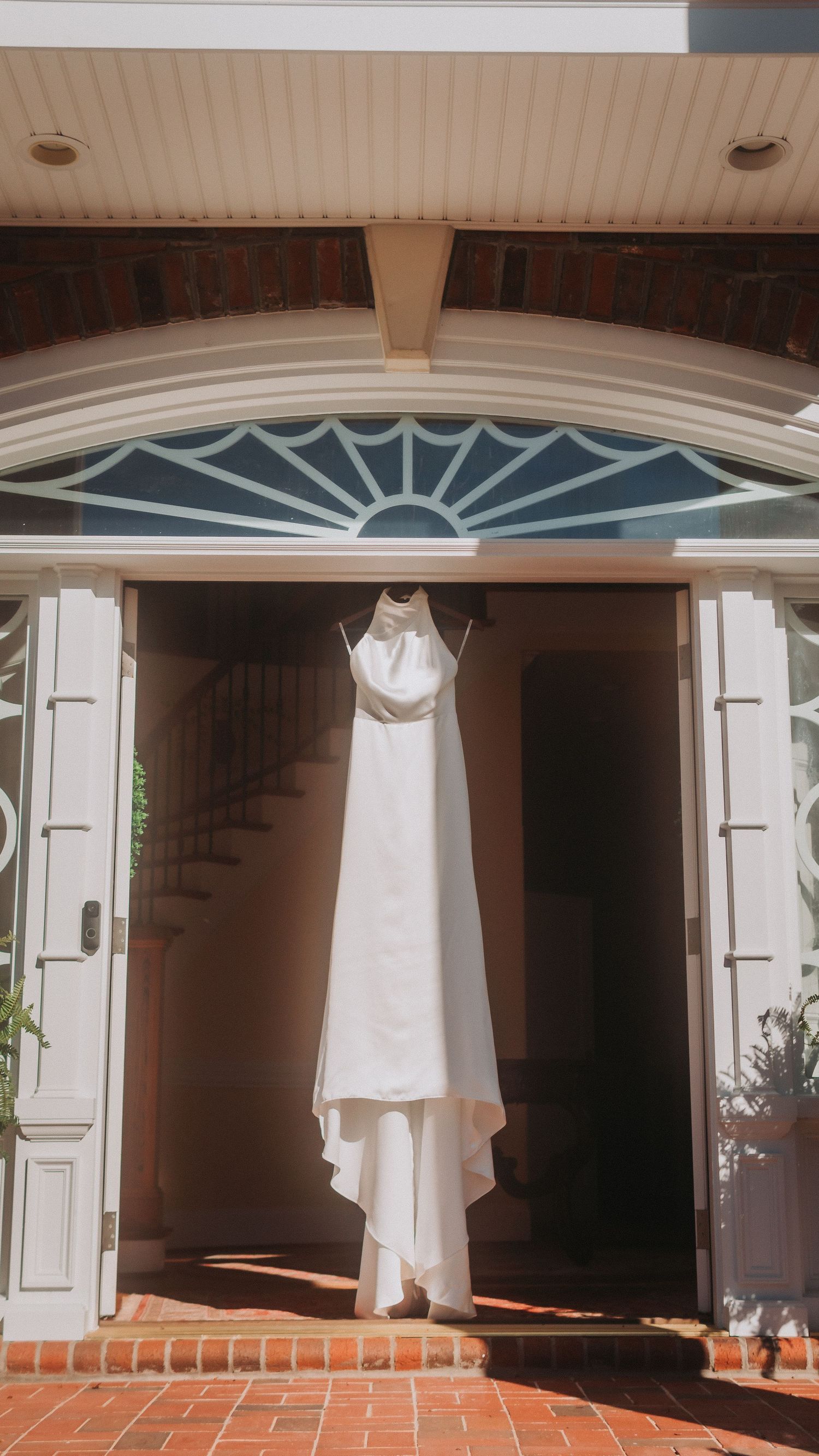White wedding dress hanging in an elegant doorway with arched window, brick flooring, and white French doors.