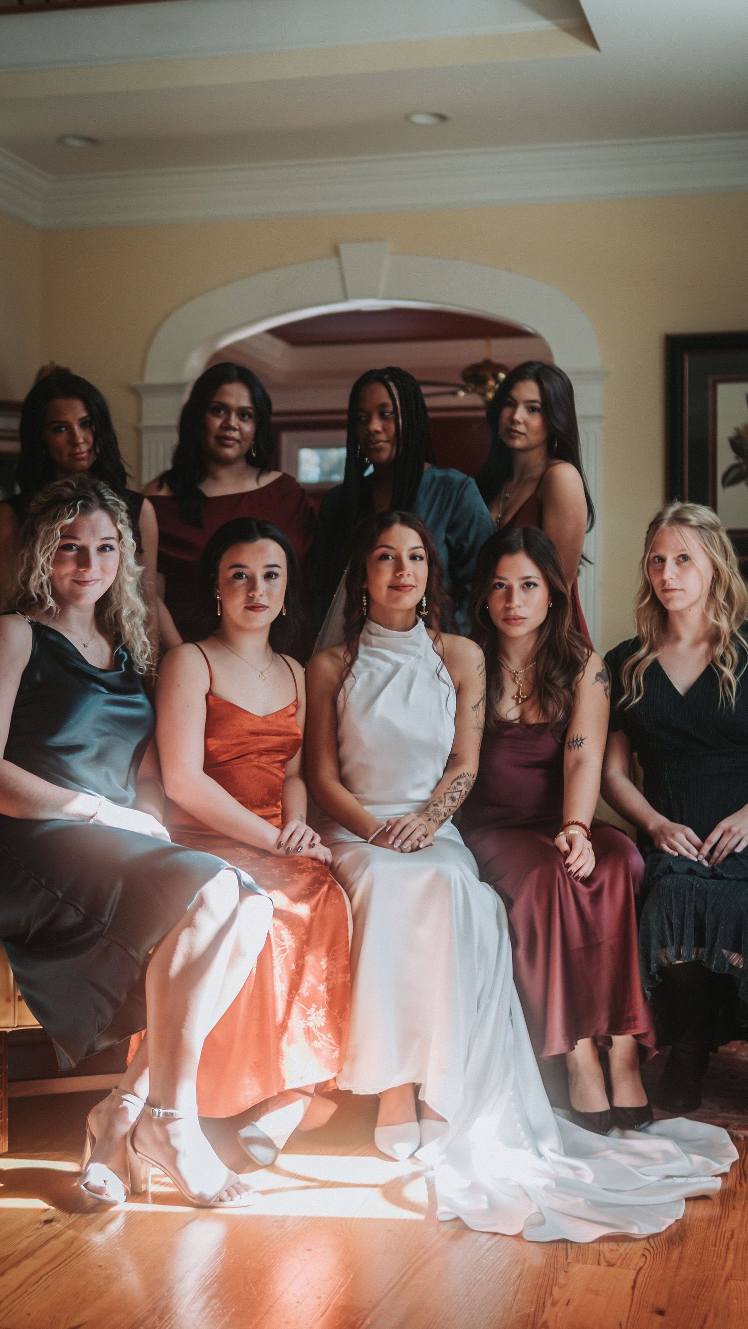 A bride in white sits with her bridesmaids in satin dresses inside an elegant home.