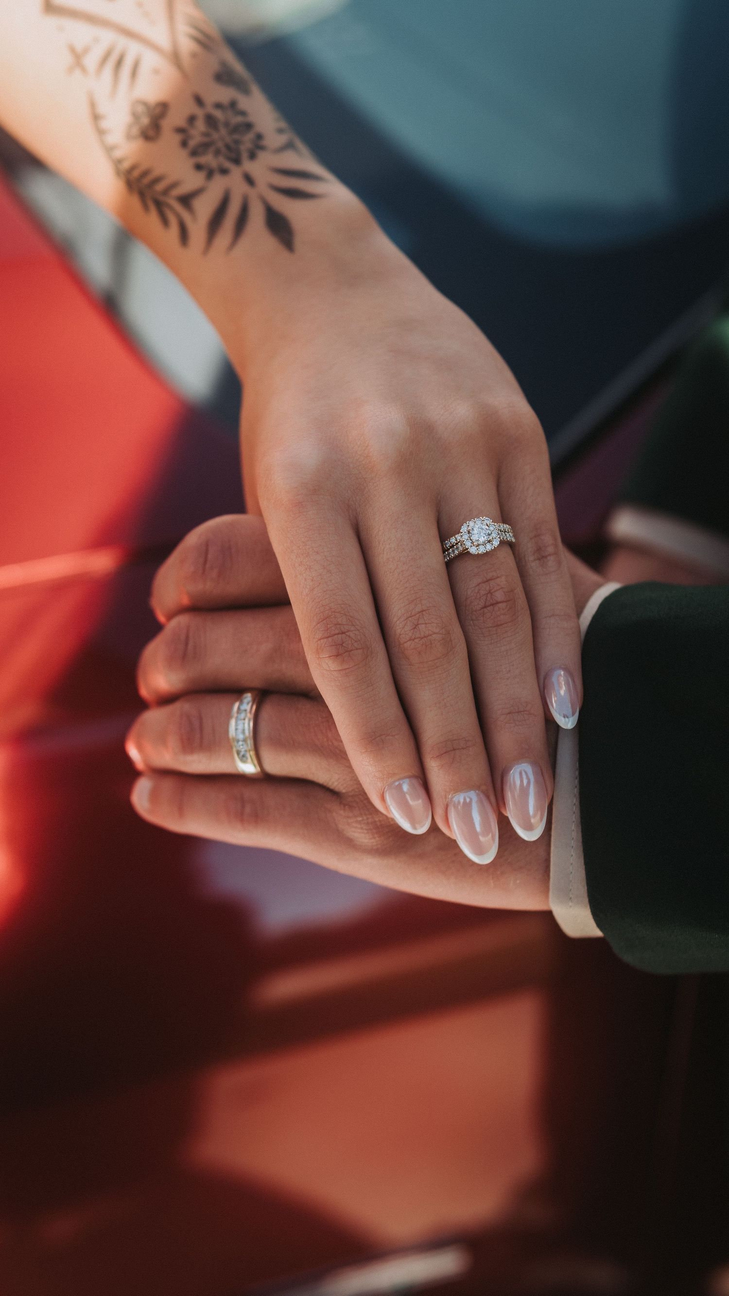 Close-up of couple's hands showing diamond engagement ring and wedding band, with floral tattoo visible on wrist.