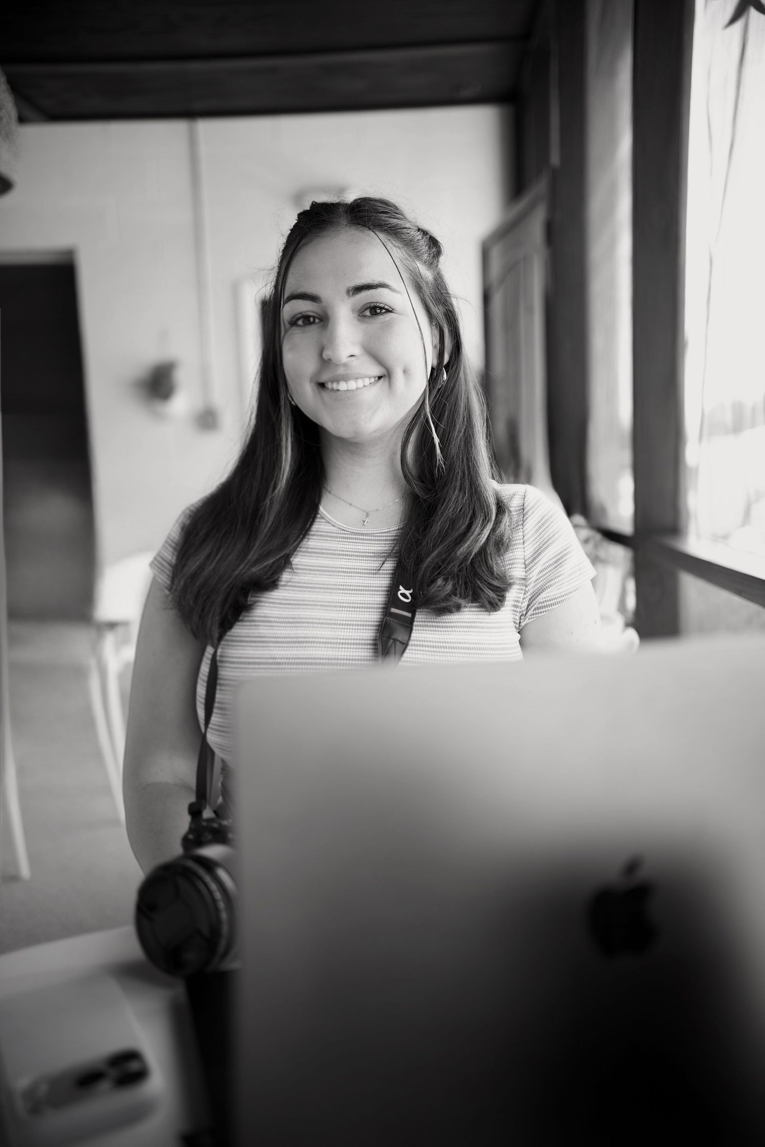 Black and white portrait of Alina, smiling CEO of Ever Joy Photography at desk with Apple laptop and headphones.