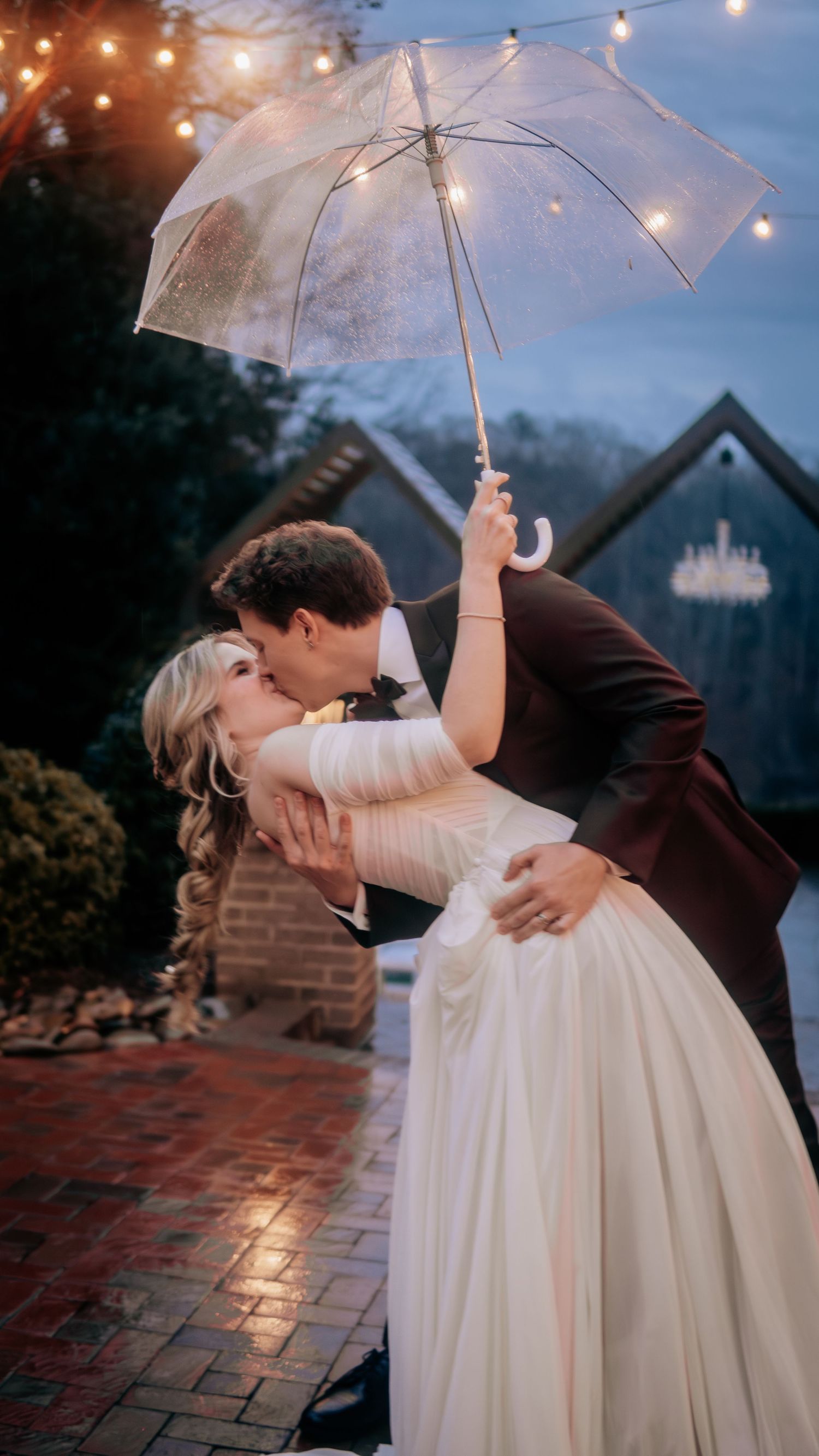 Bride and groom share a romantic dip kiss under a clear umbrella on a brick path at dusk during their wedding.
