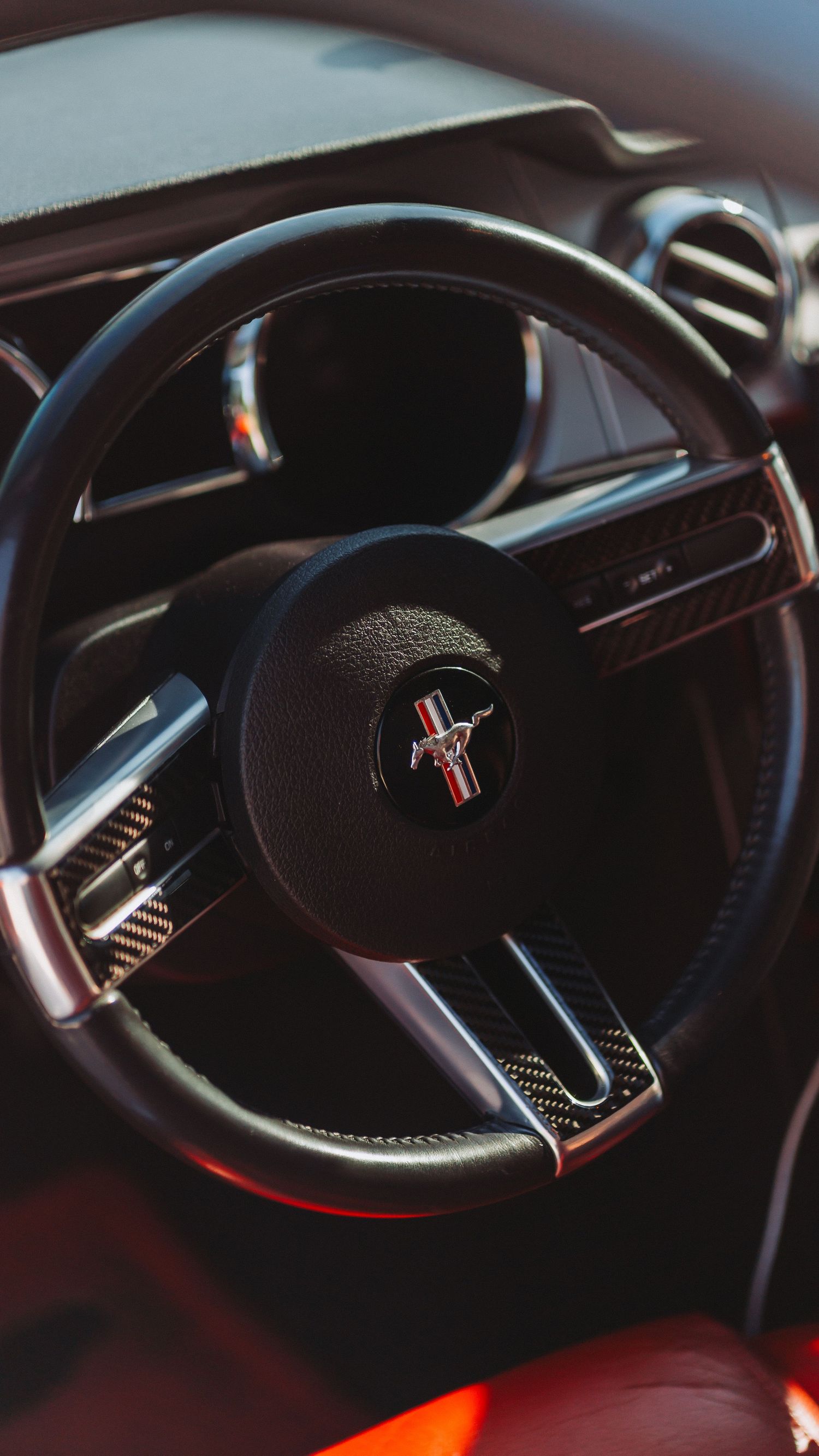 Close-up of a Ford Mustang steering wheel with iconic running horse logo and carbon fiber trim accents.
