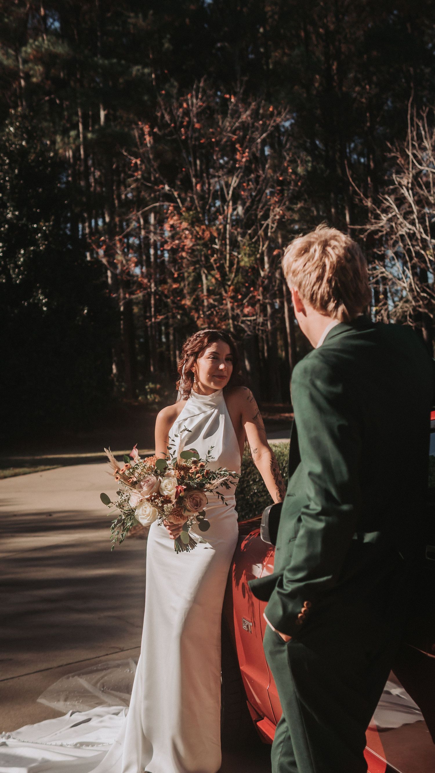 Bride in white halter gown holding bouquet stands by red car with groom in green suit amid sunlit trees.