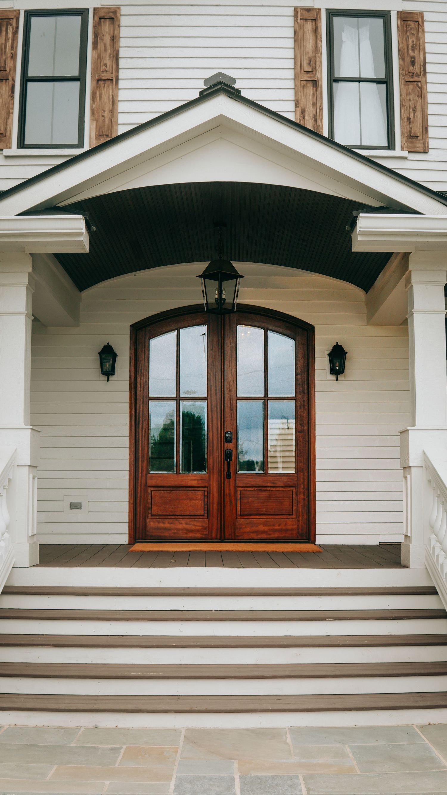 Elegant white home entrance with arched mahogany double doors, black lanterns, covered portico, and stone steps.
