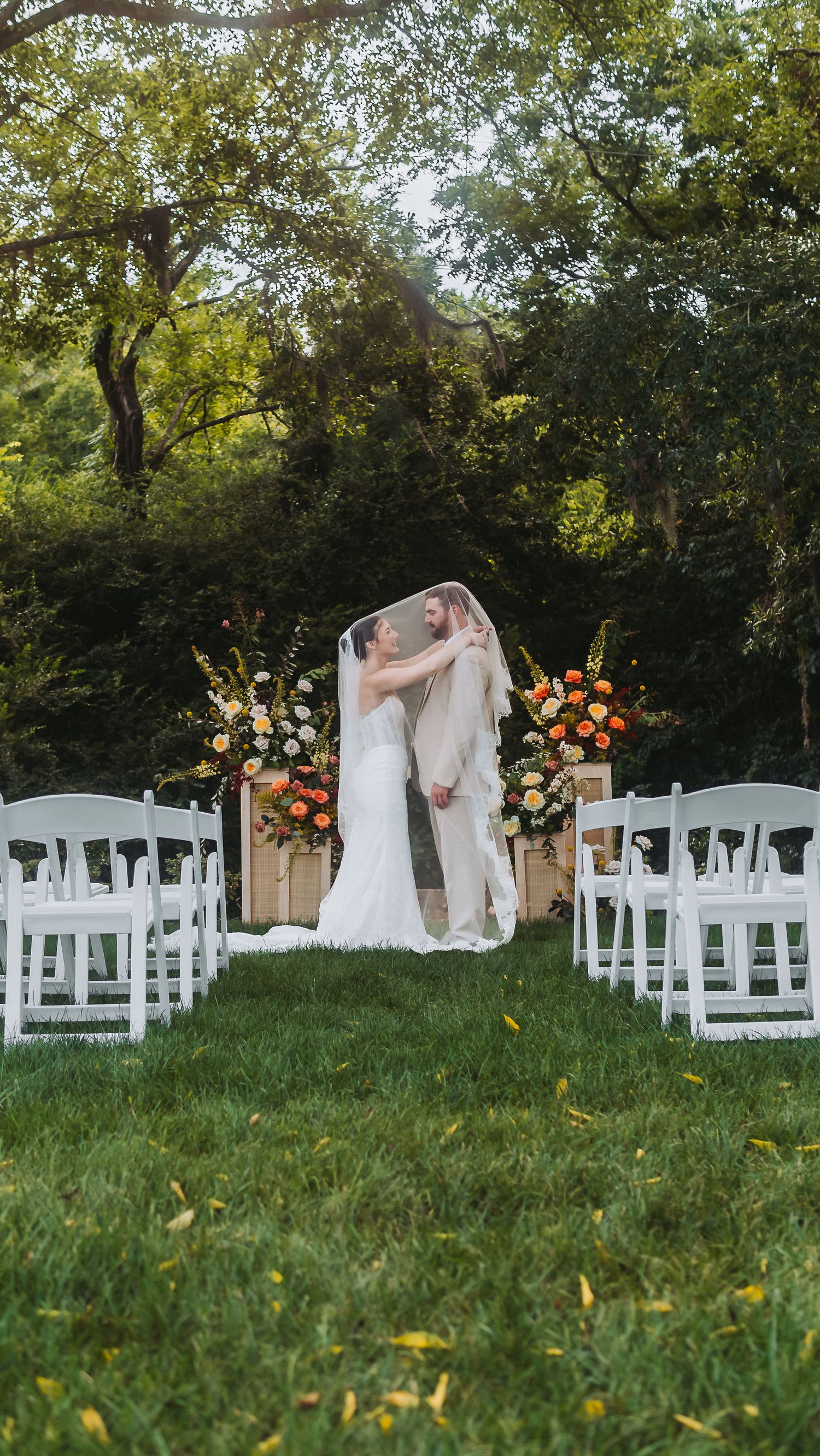 Two brides share a first kiss under a veil at an outdoor wedding ceremony with white chairs and floral arrangements.
