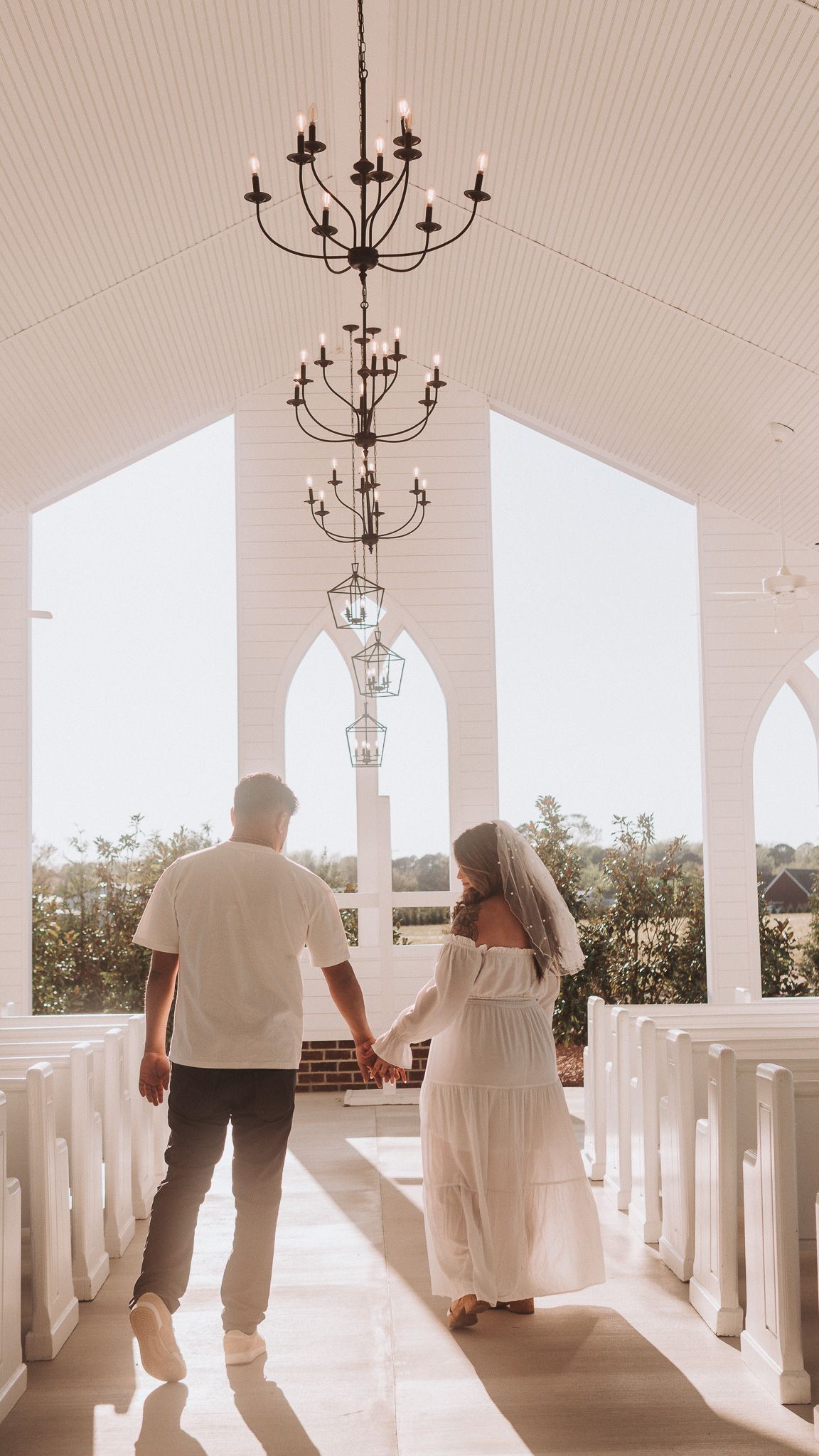 Couple holding hands walking down the aisle of a white chapel with chandeliers and arched windows.