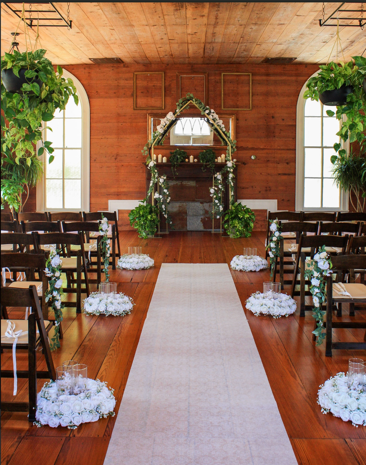Elegant wedding ceremony aisle with white floral arrangements, wooden chairs, greenery arch, and rustic wood-paneled chapel interior.