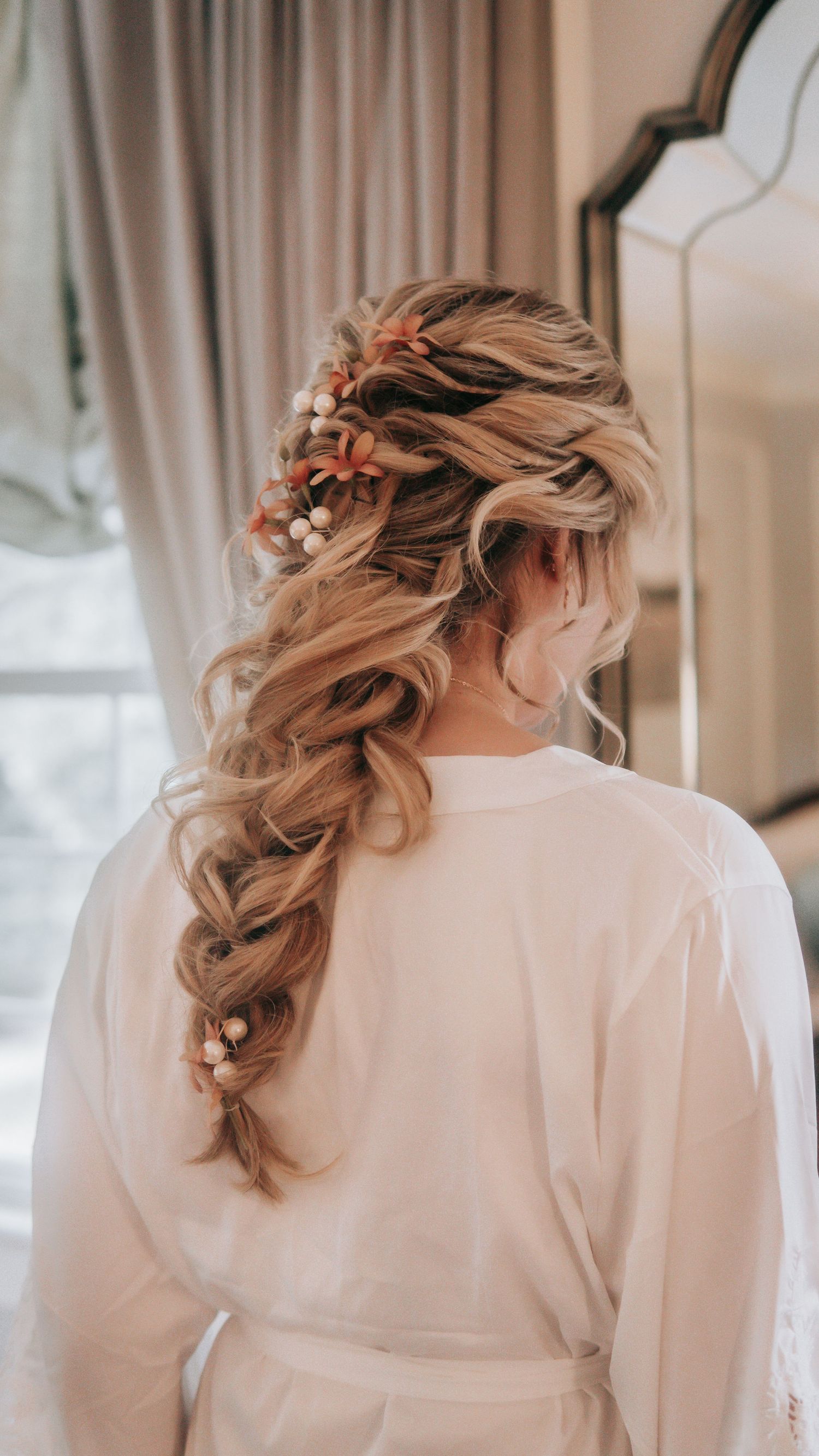 Bride with elegant braided hairstyle adorned with small floral pins, wearing a white robe, viewed from behind.