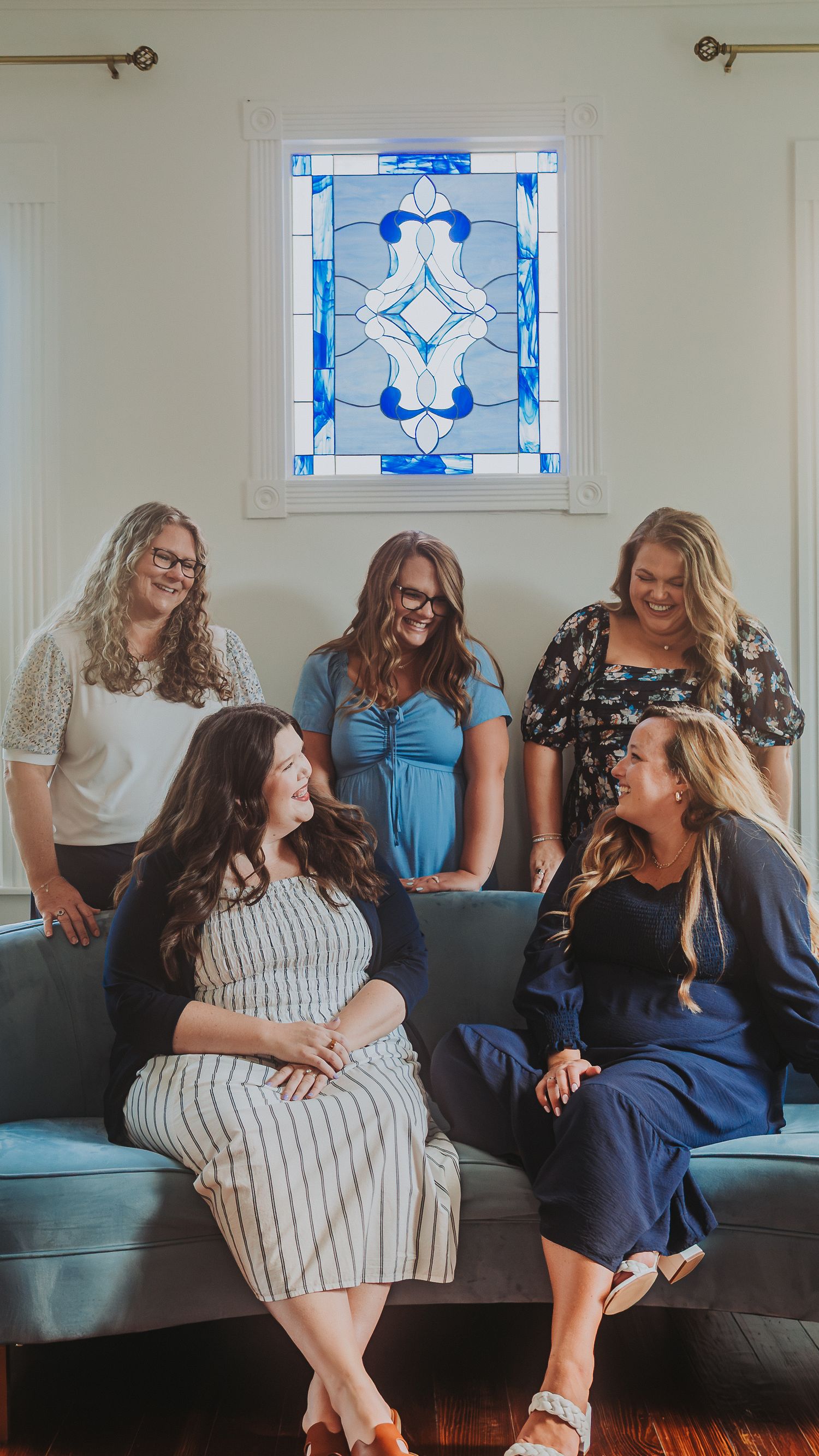 Five women smiling and laughing together in a bright room with a blue stained glass window above a gray sofa.