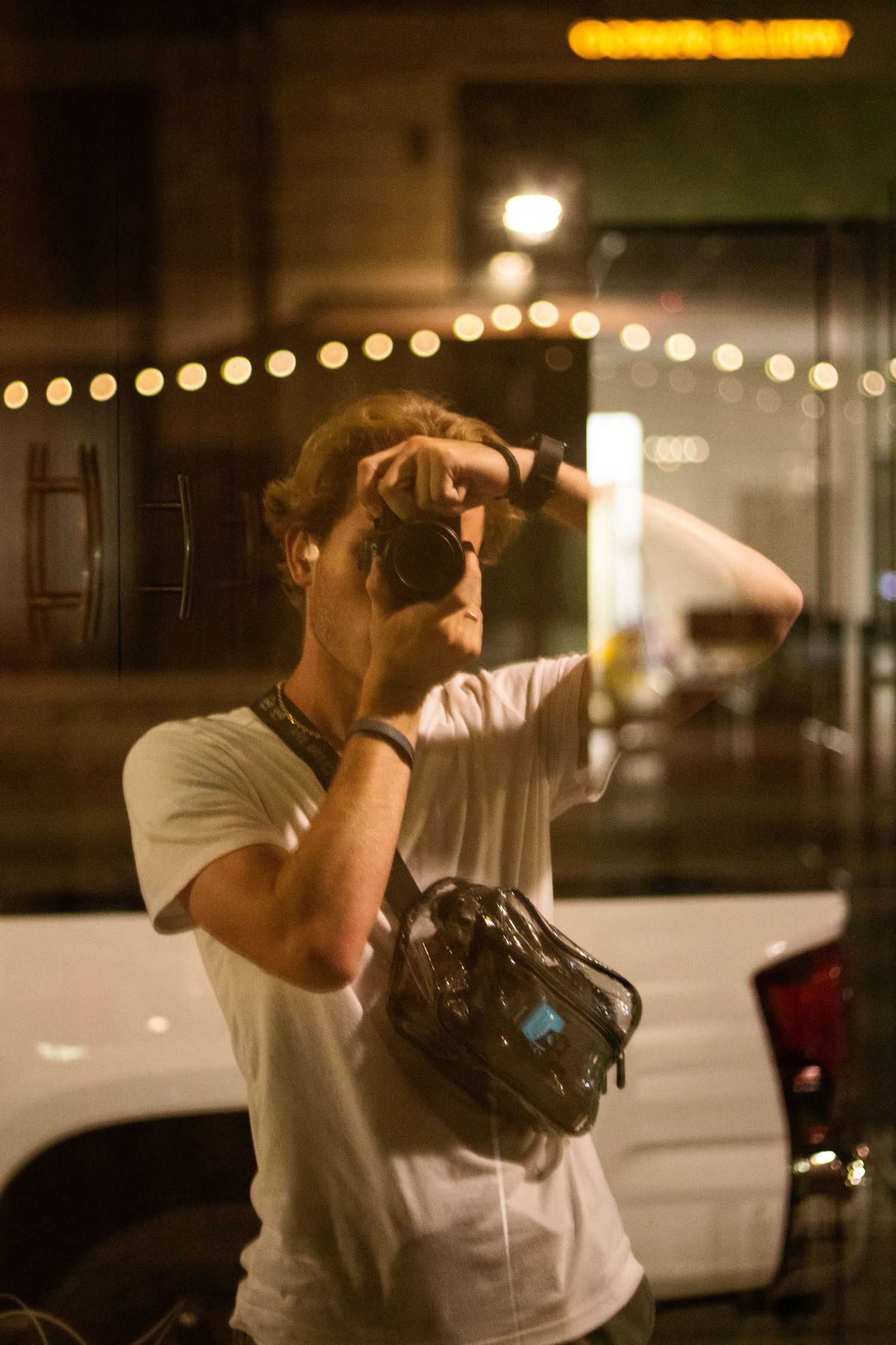 Tyler talking a photo of his reflection in a window, lights are strung up in the background
