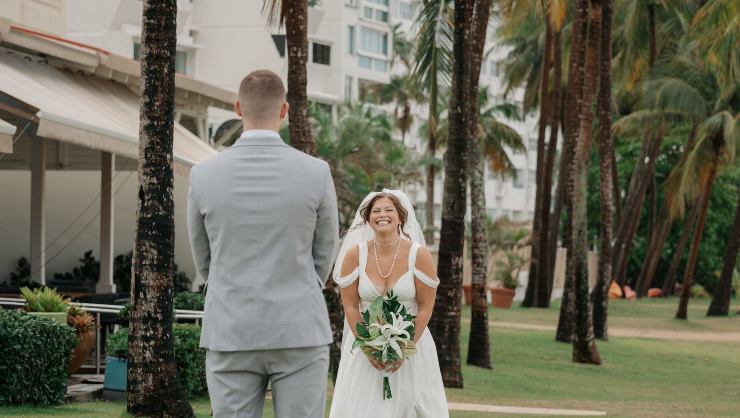 Bride in white gown holding bouquet walks toward groom in gray suit among tropical palm trees at outdoor wedding venue.