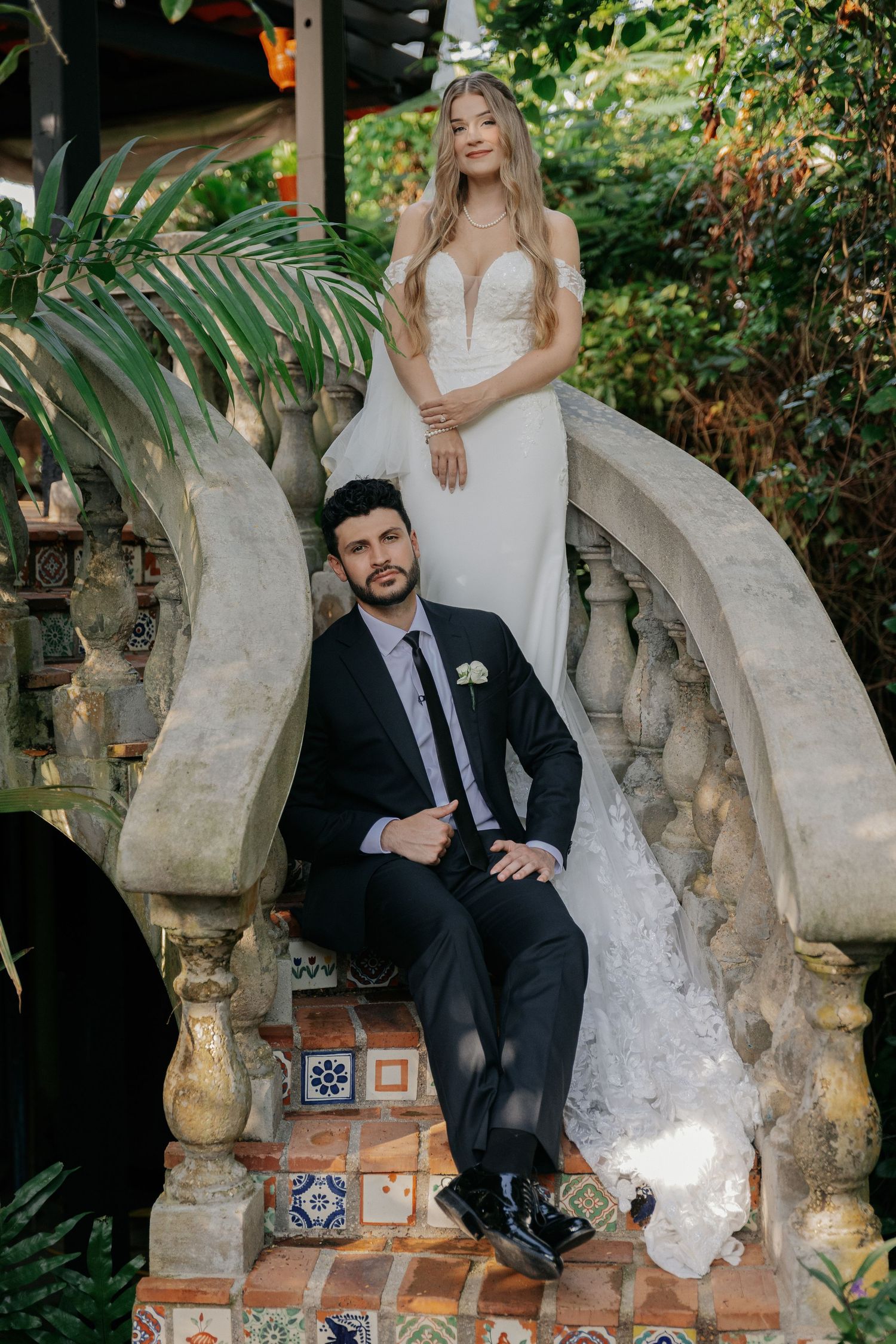 Bride in white gown and groom in black suit pose on ornate stone staircase surrounded by lush greenery.