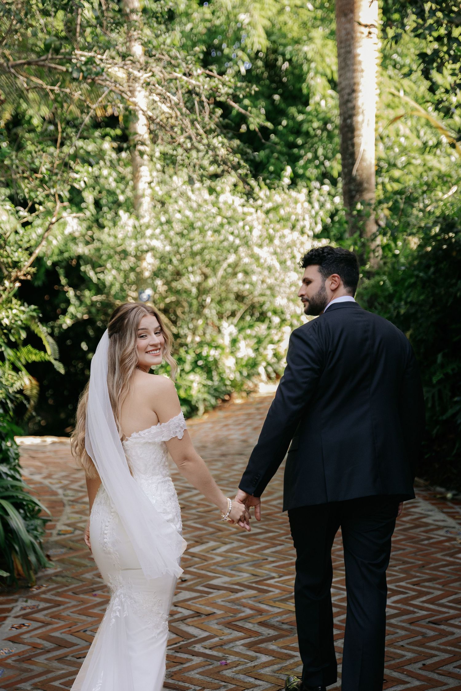 Bride in off-shoulder lace gown and veil holds hands with groom in black suit walking through lush garden path.