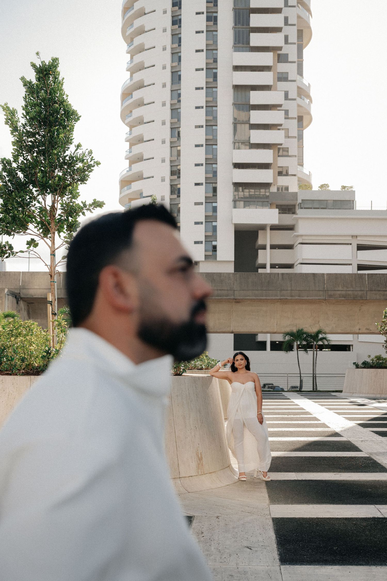 Bearded man in white shirt stands in foreground as woman in white dress poses near modern high-rise building outdoors.
