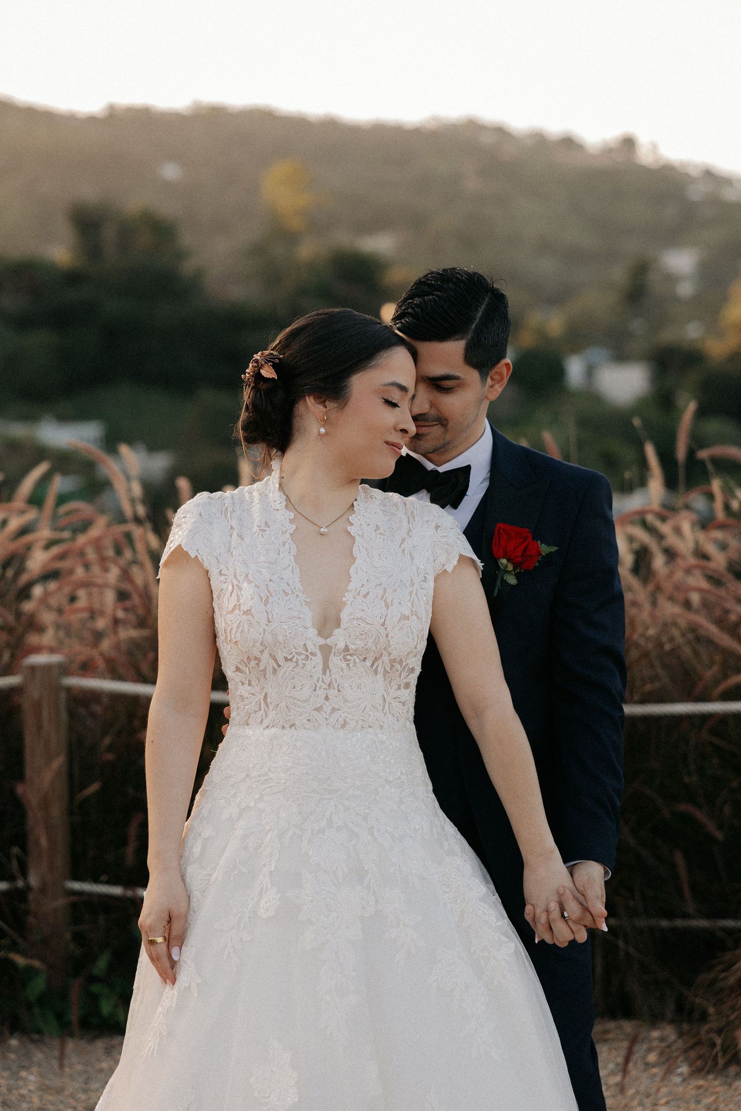 Bride in lace gown and groom in navy tuxedo share a romantic moment at golden hour with hills in the background.