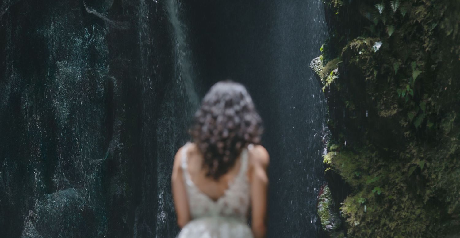 A person with curly hair in a formal dress stands facing a dark waterfall against a backdrop of greenery.