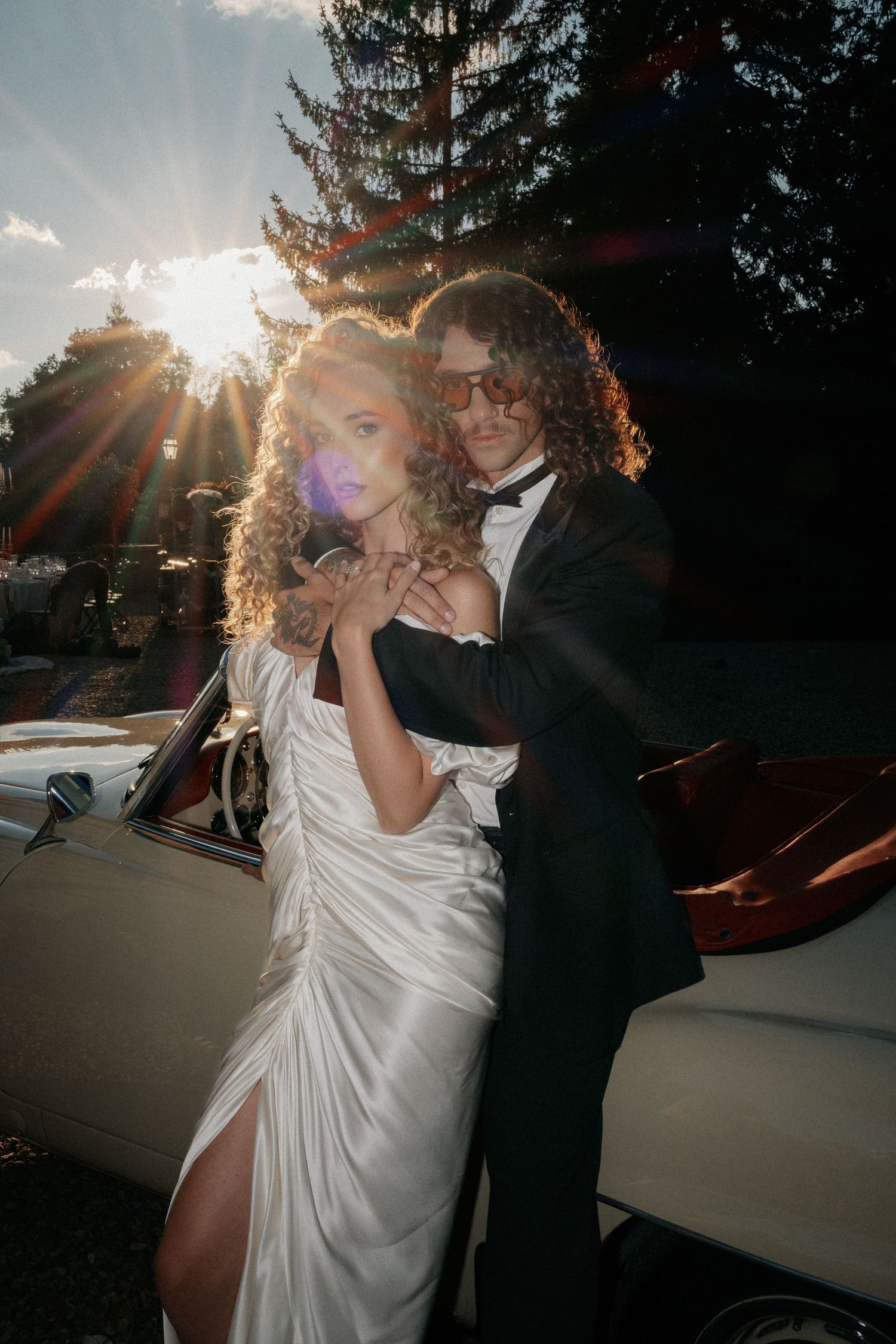 Elegant couple in formal wedding attire posing by a vintage convertible car at sunset with sun flare.