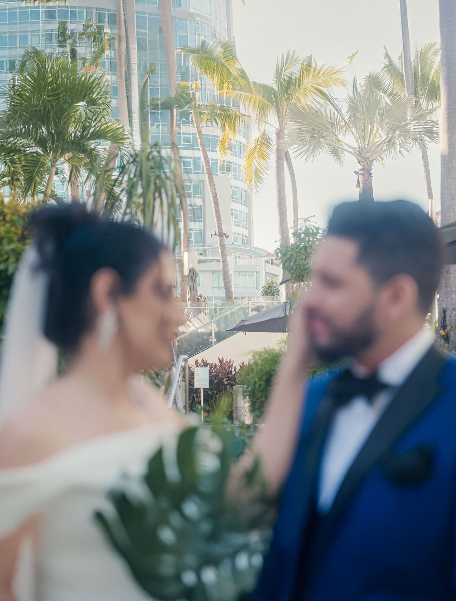 Wedding couple in tropical garden setting with palm trees and high-rise building in background during sunset ceremony.