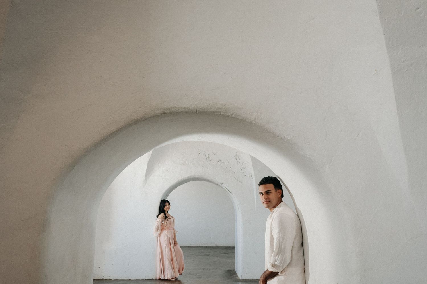 Couple in white arched hallway, woman in pink dress, man in white shirt, romantic engagement photo.