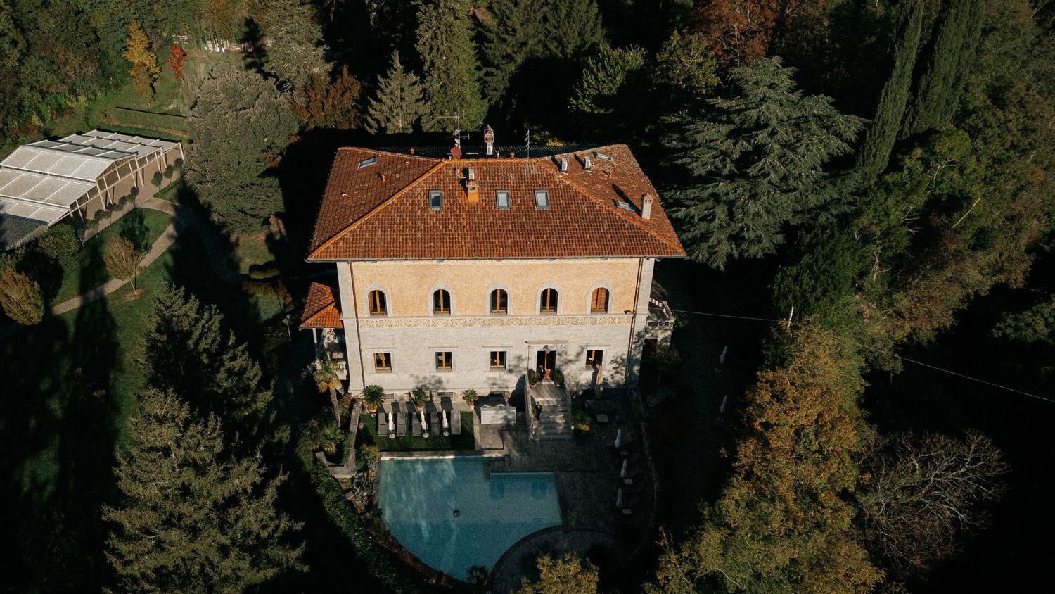 Aerial view of a historic Italian villa with terracotta roof, pool, and lush autumn forest surroundings.