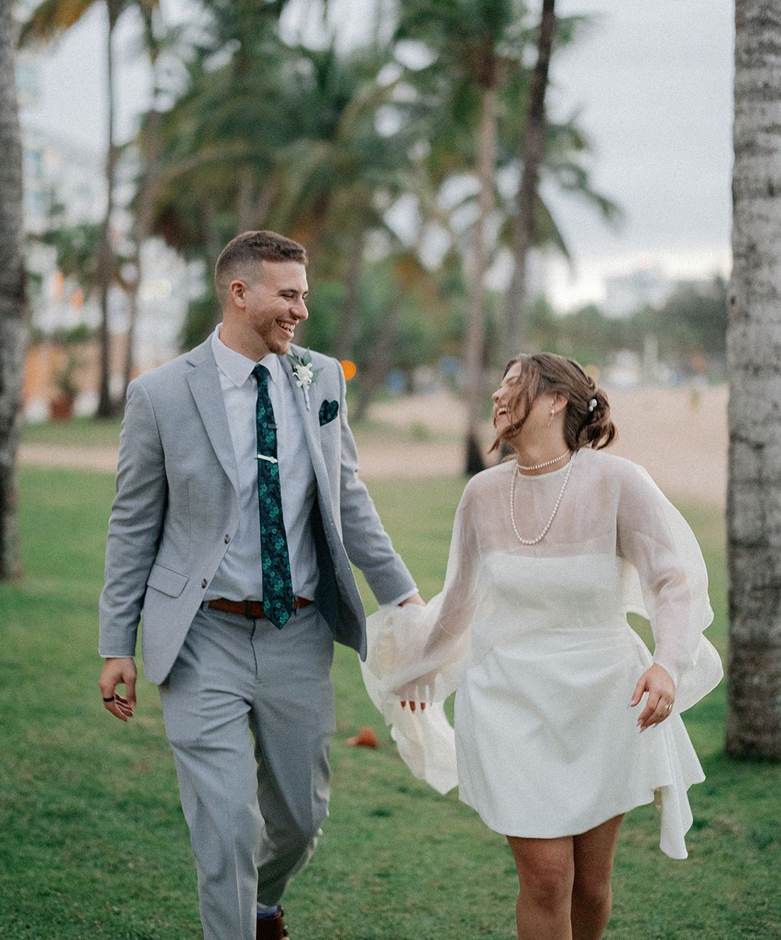 Newlywed couple walking hand-in-hand on tropical beach lawn, bride in white mini dress, groom in gray suit with green tie.