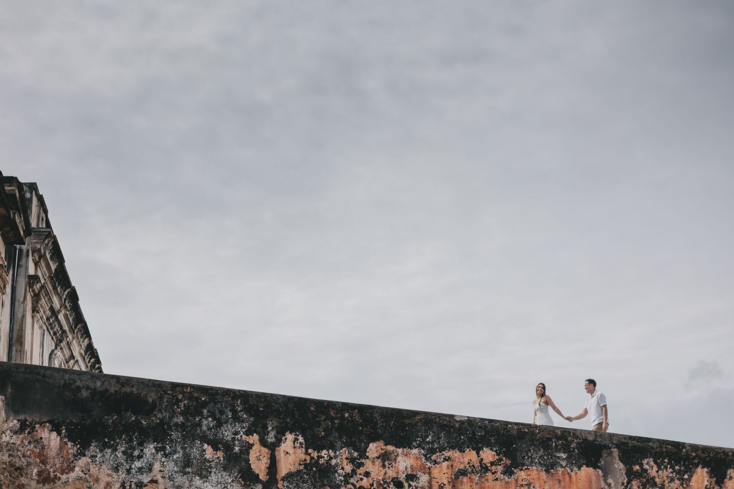 Couple holding hands on top of an aged stone wall with a historic building and overcast sky backdrop.