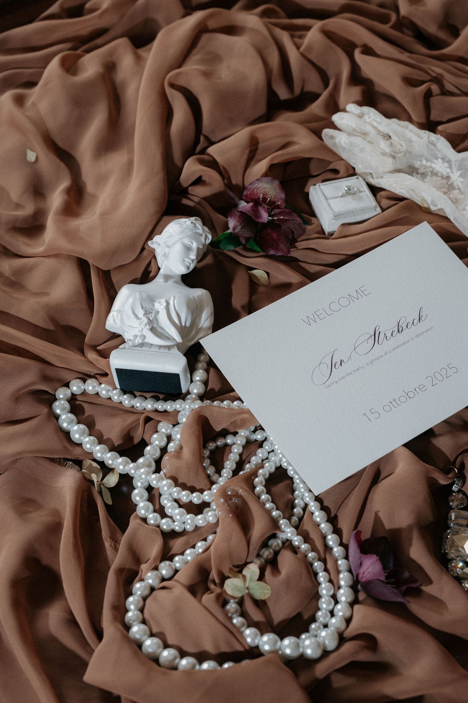 Elegant wedding flat lay with pearl necklace, white bust, lace gloves, ring box, and welcome card on brown silk fabric.