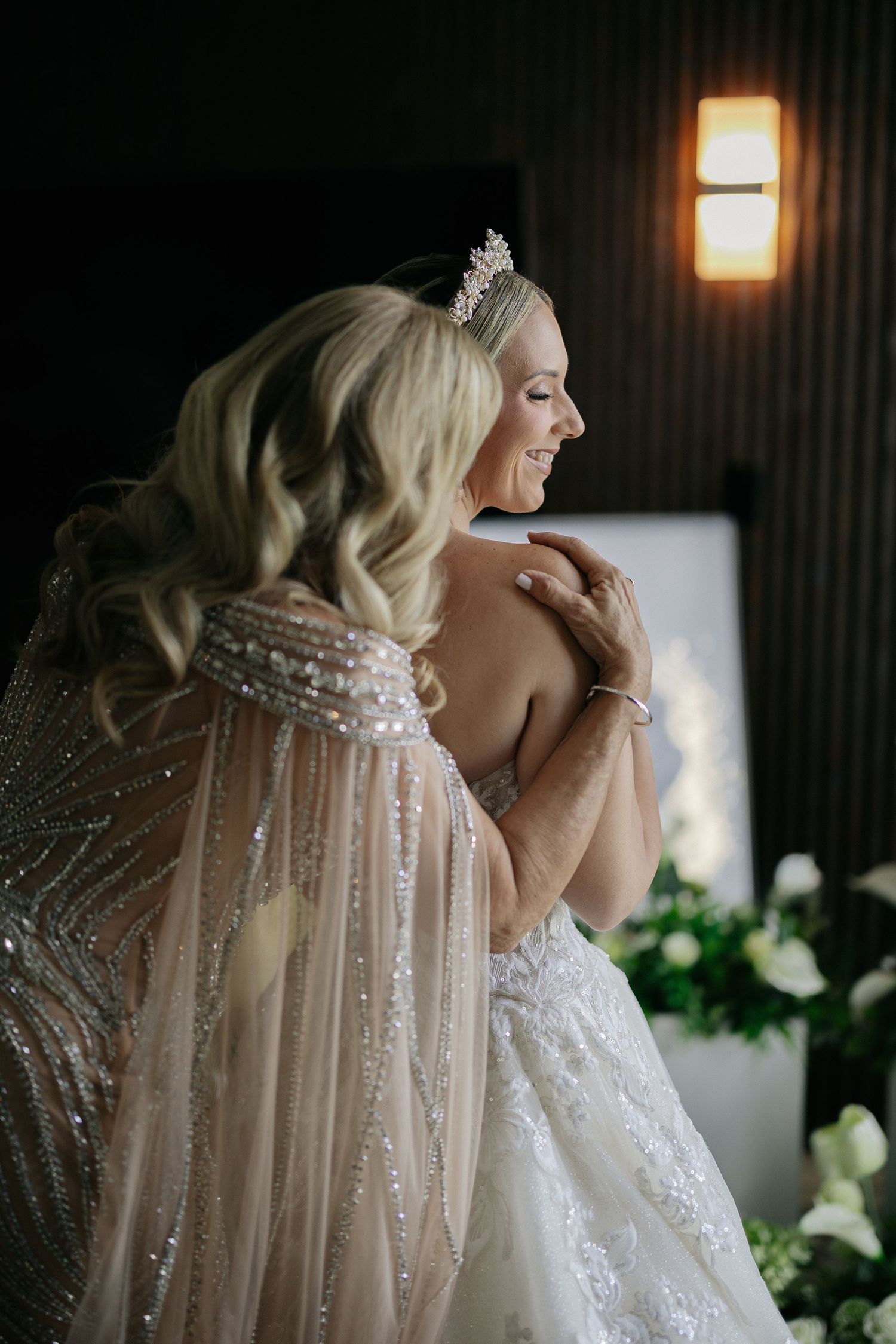Bride in a lace gown and tiara smiles as a woman in a beaded cape adjusts her dress in a softly lit room.