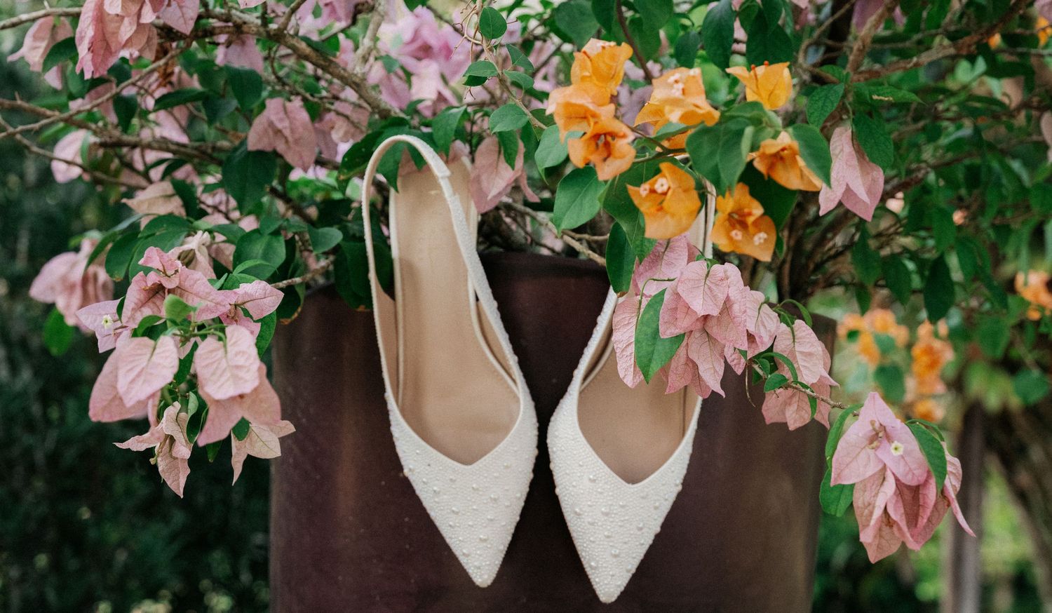 White pearl bridal heels hanging against vibrant pink and orange bougainvillea flowers on a rustic brown planter.