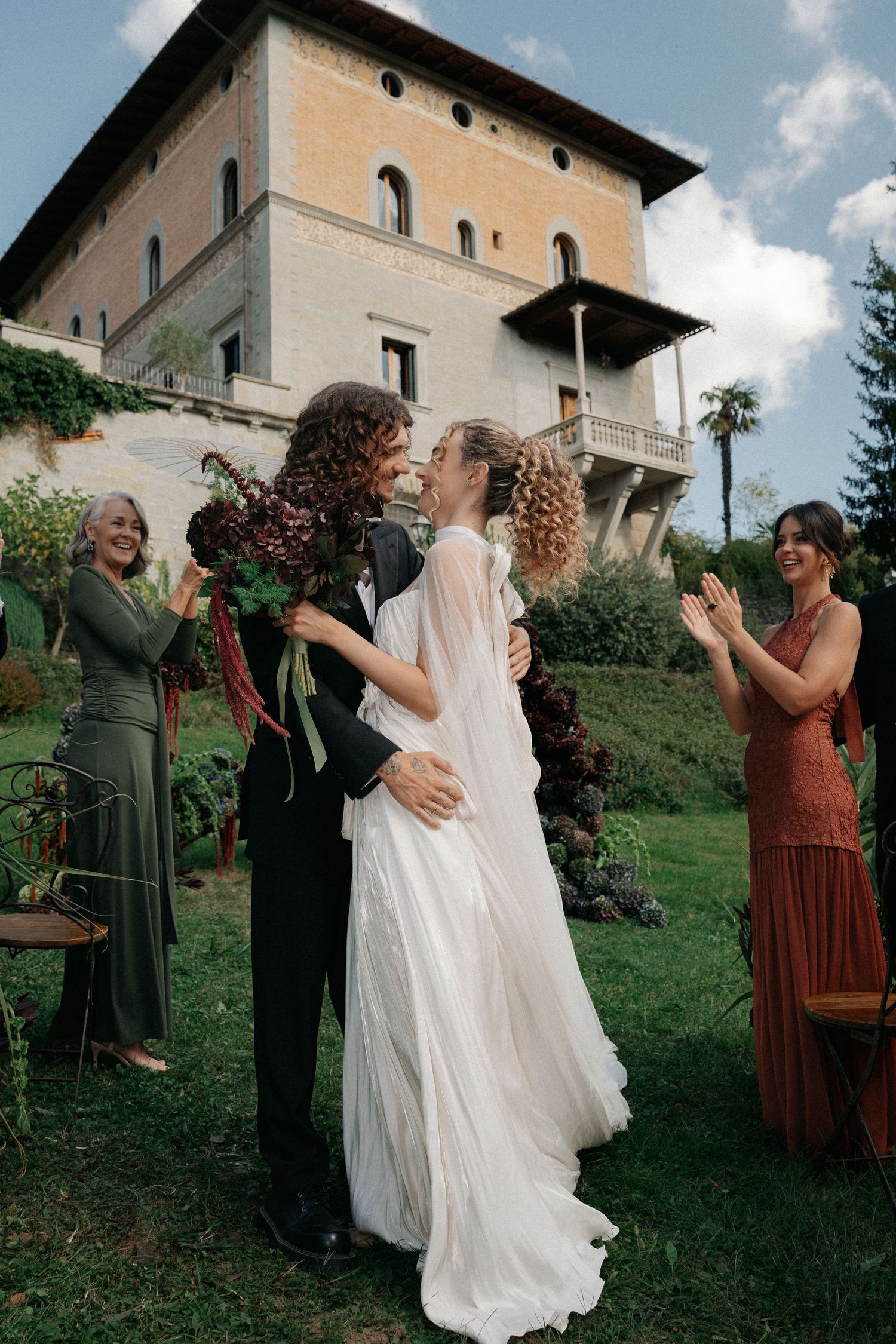 Bride and groom share first kiss outdoors at elegant Italian villa wedding ceremony with guests applauding.